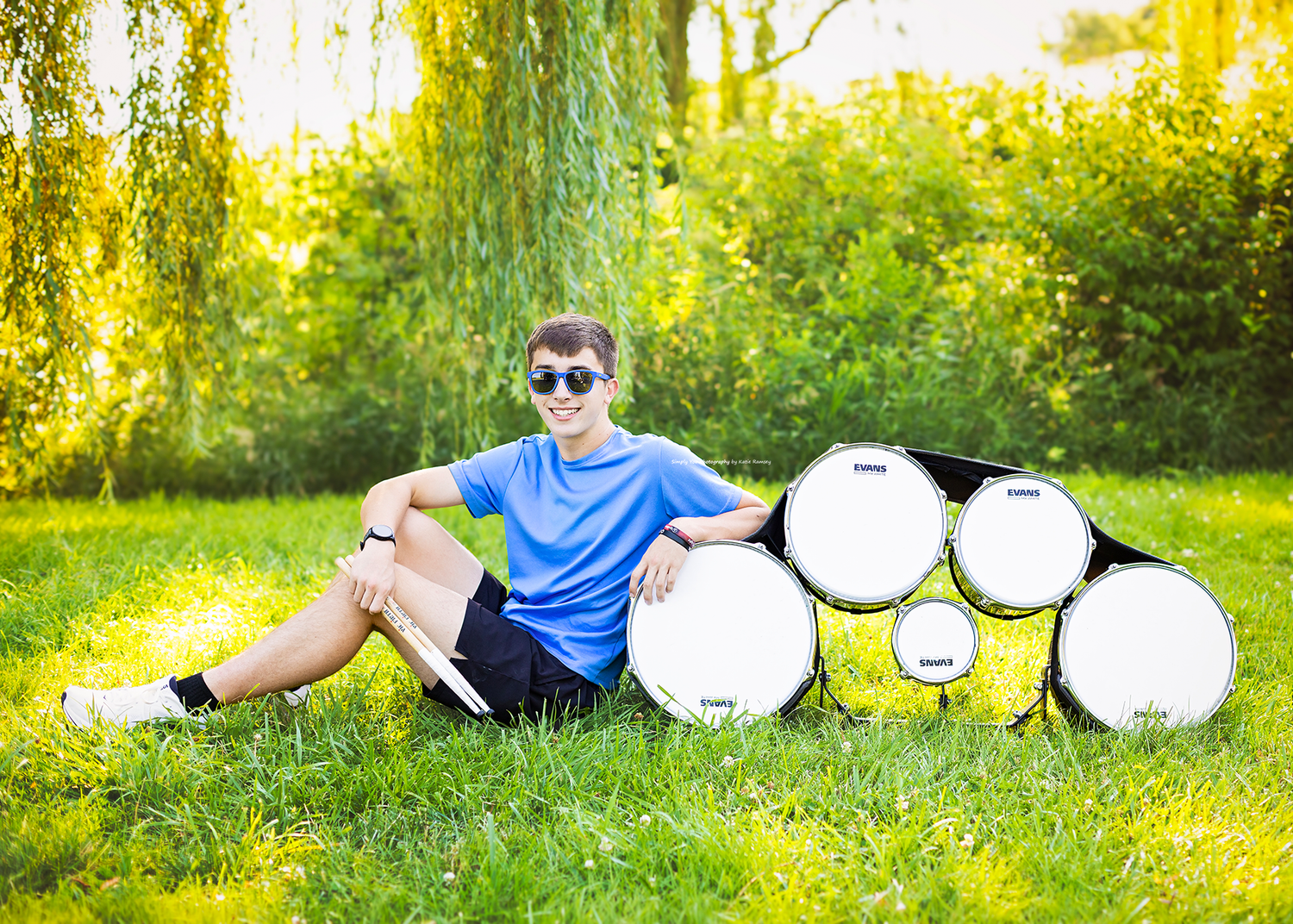 Boy in blue shirt and sunglasses sitting on grass leaning against a stack of white drums outdoors.