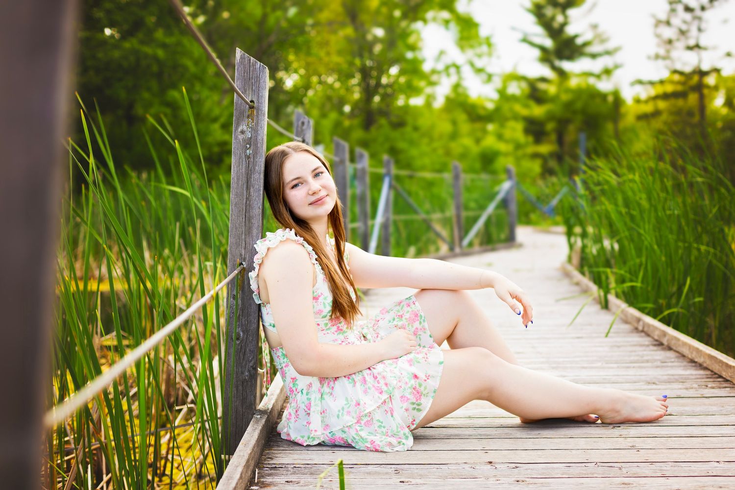 Young woman in floral dress sitting on wooden boardwalk surrounded by lush green reeds and trees.
