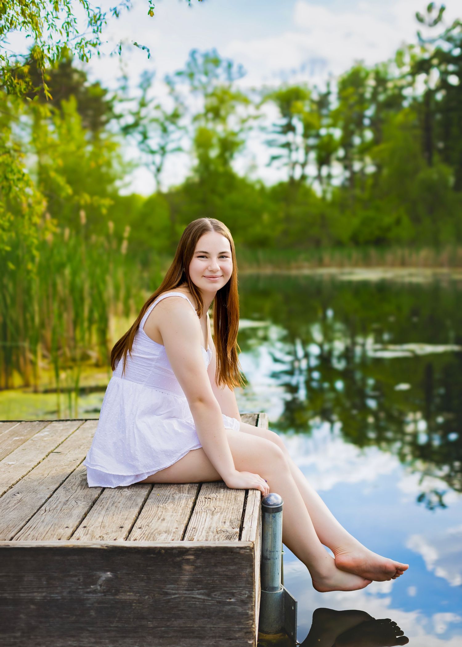 Young woman in white dress sitting barefoot on a wooden dock by a serene reflective lake.