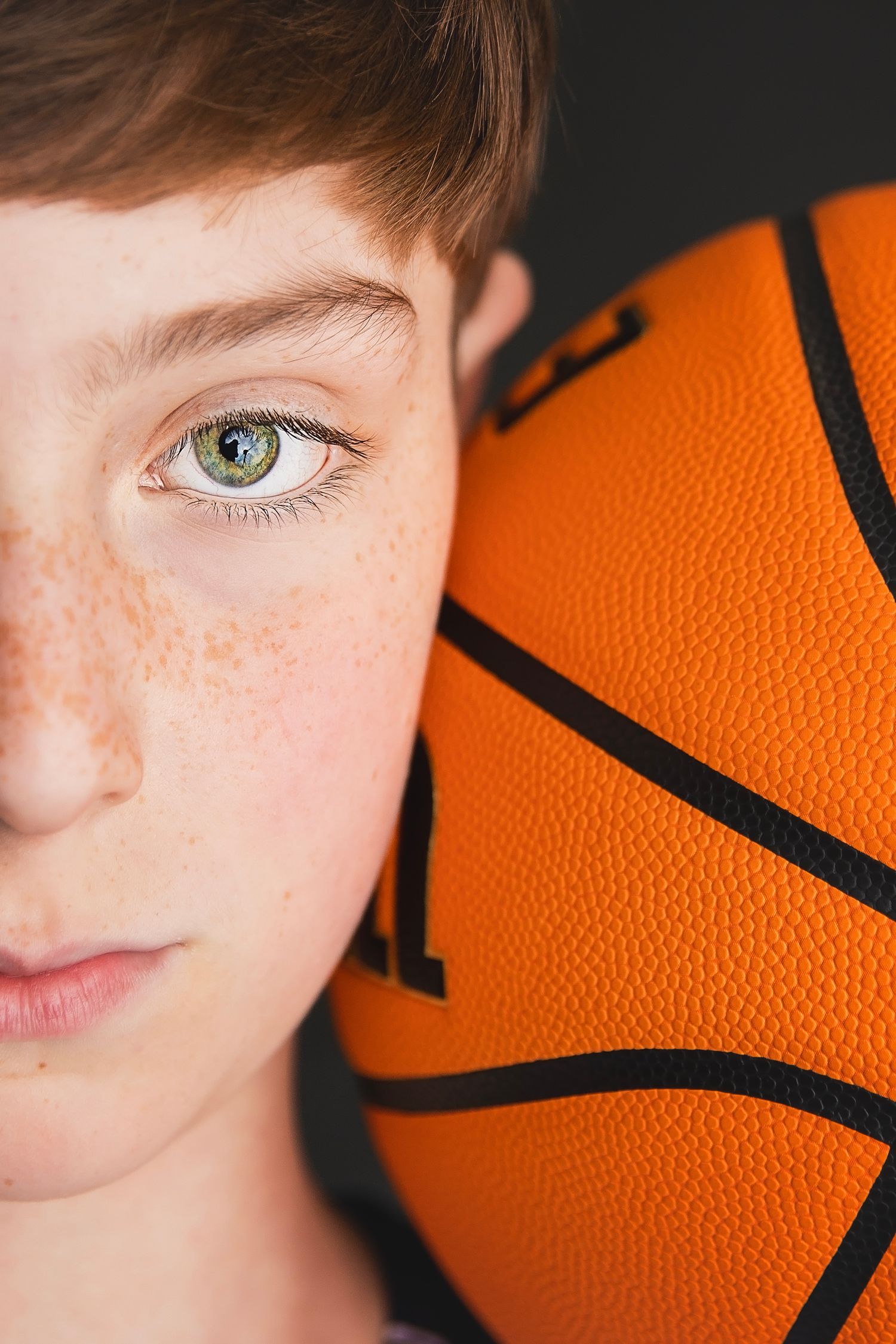 Close-up of a freckled boy with green eyes with a basketball pressed against his face.
