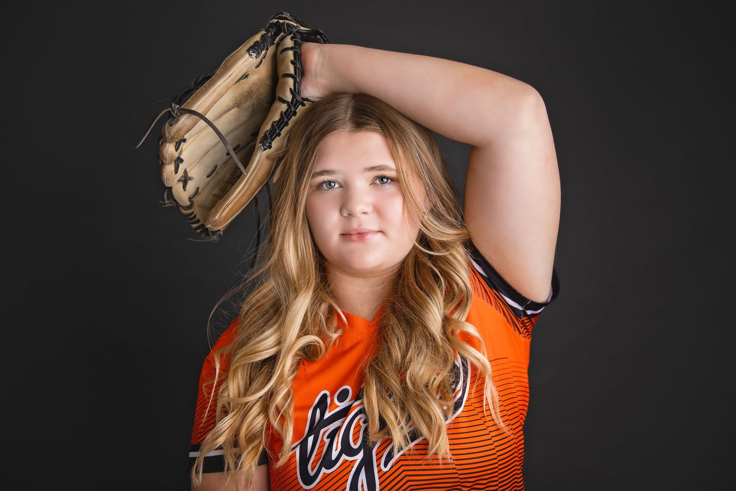 Young softball player in orange Tigers uniform holds glove above head in dramatic studio pose.