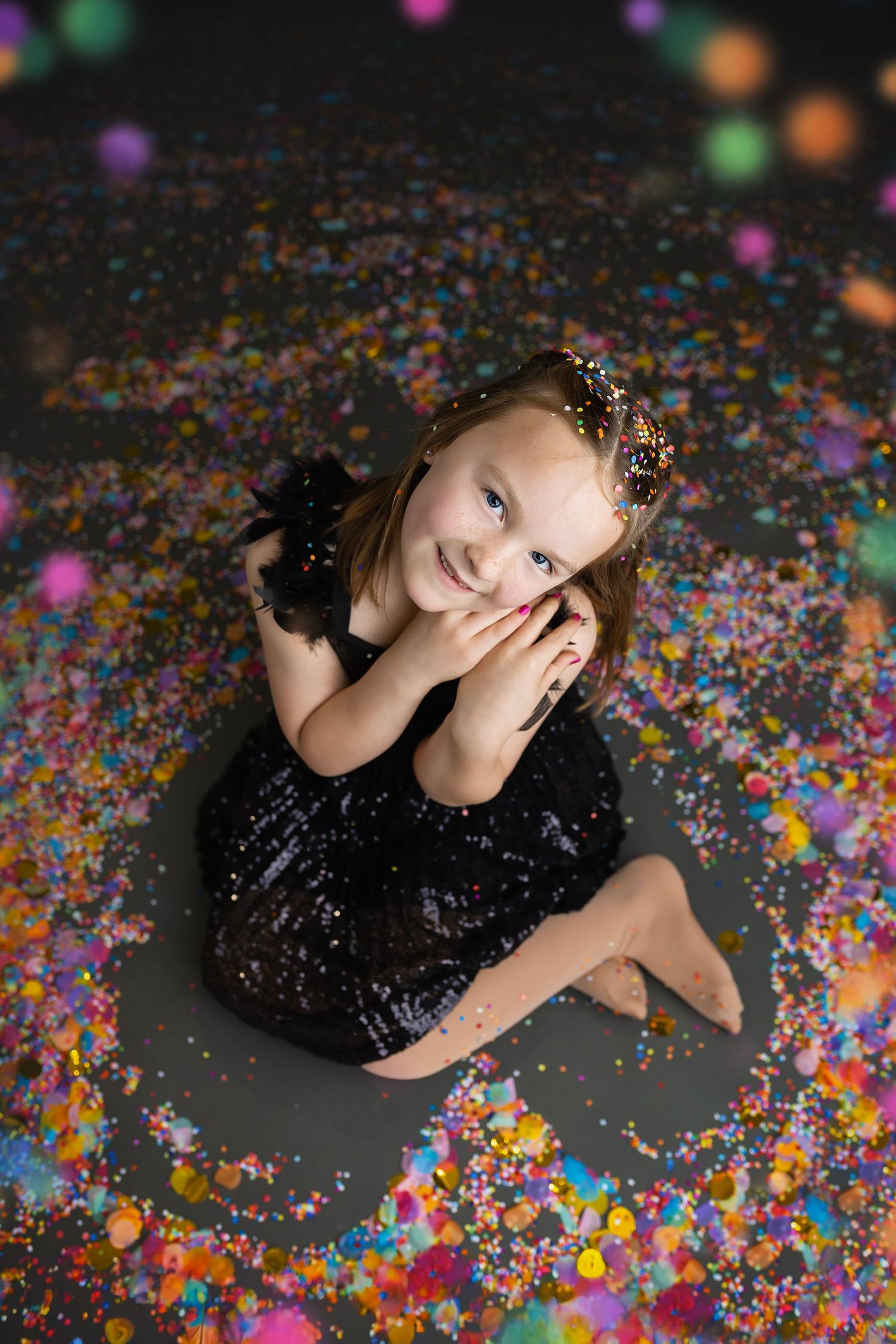 Young girl in black sequin dress sitting on floor surrounded by colorful confetti.