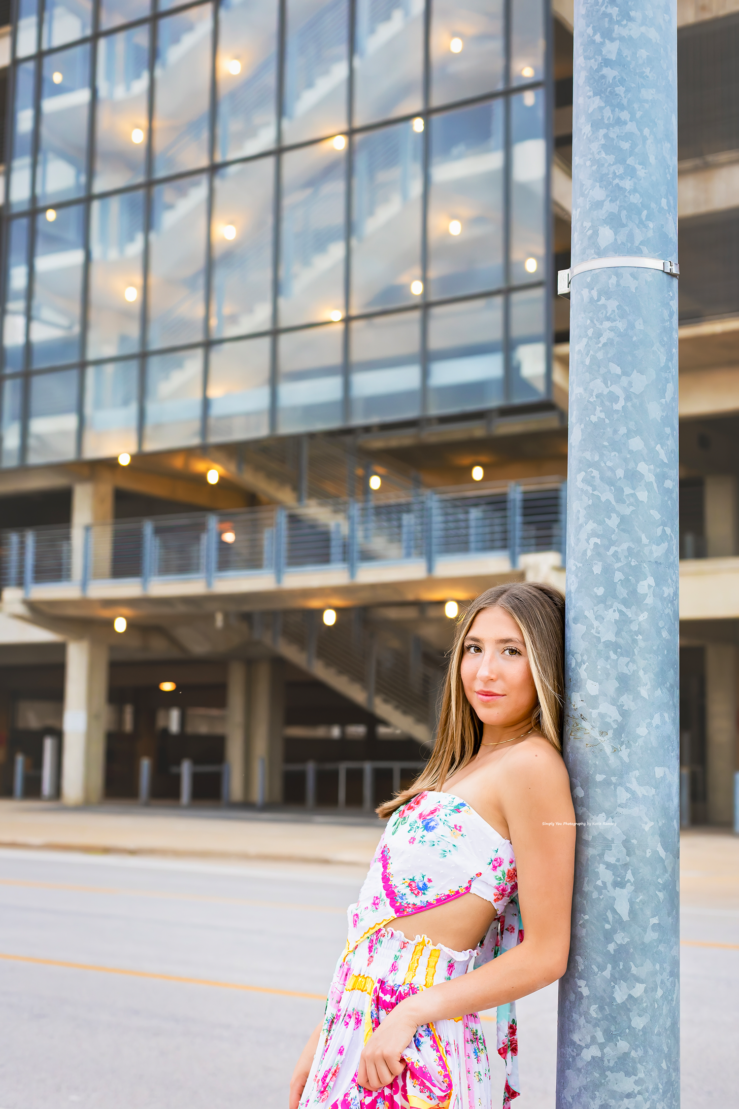 Young woman in floral maxi dress leaning against urban pole with modern glass building backdrop.