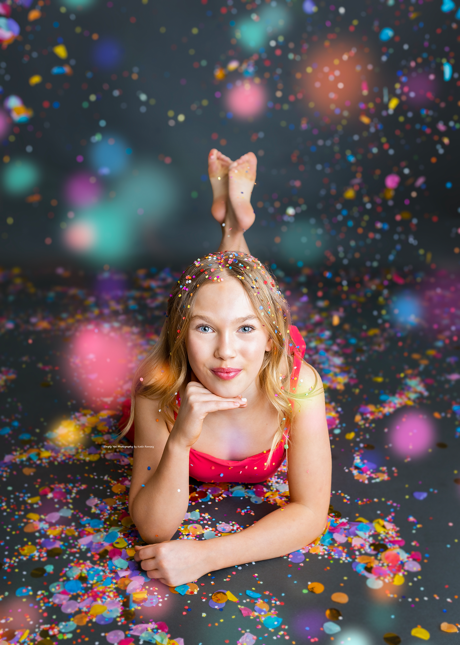 Girl in a red dress posing on the floor surrounded by vibrant falling confetti in a studio.