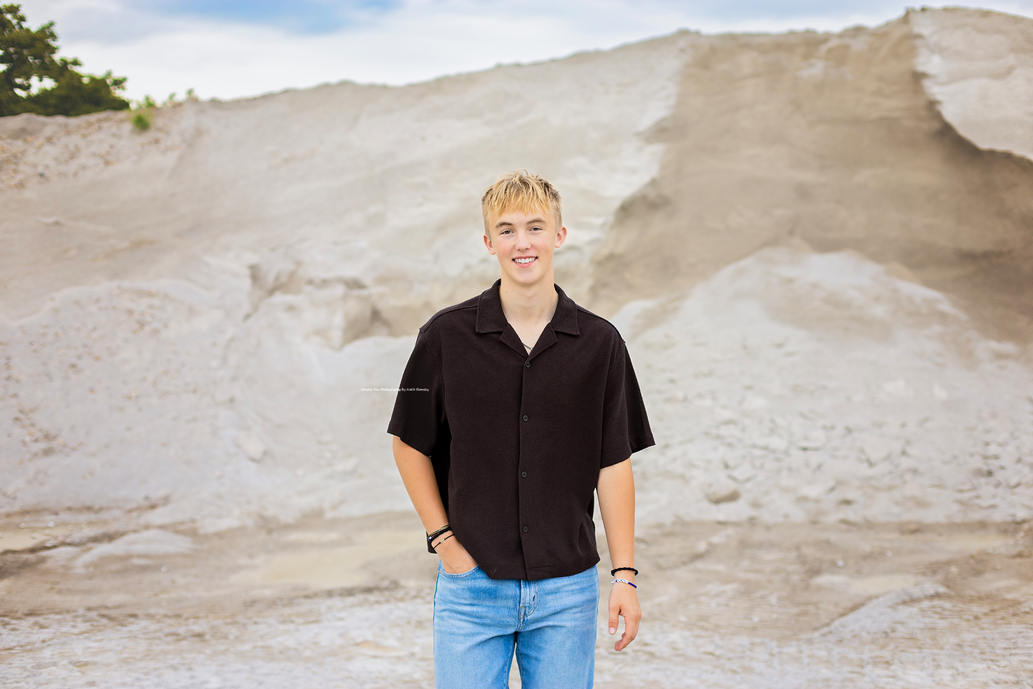 Young blonde teen boy smiling in black shirt and jeans, posing outdoors near white sandy rock formations.