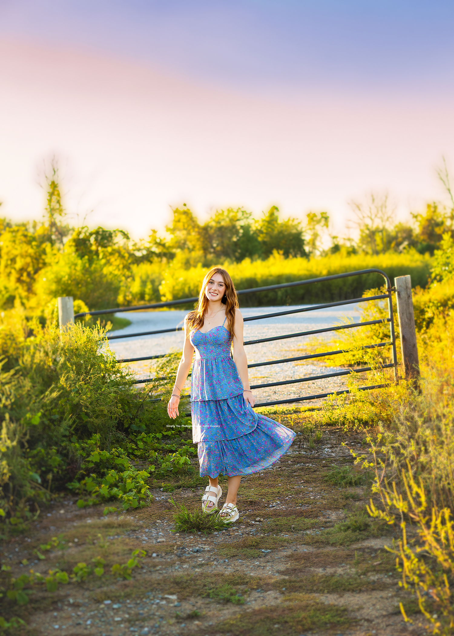 Young woman in blue floral dress walking on a sunlit path by a wooden fence at golden hour.