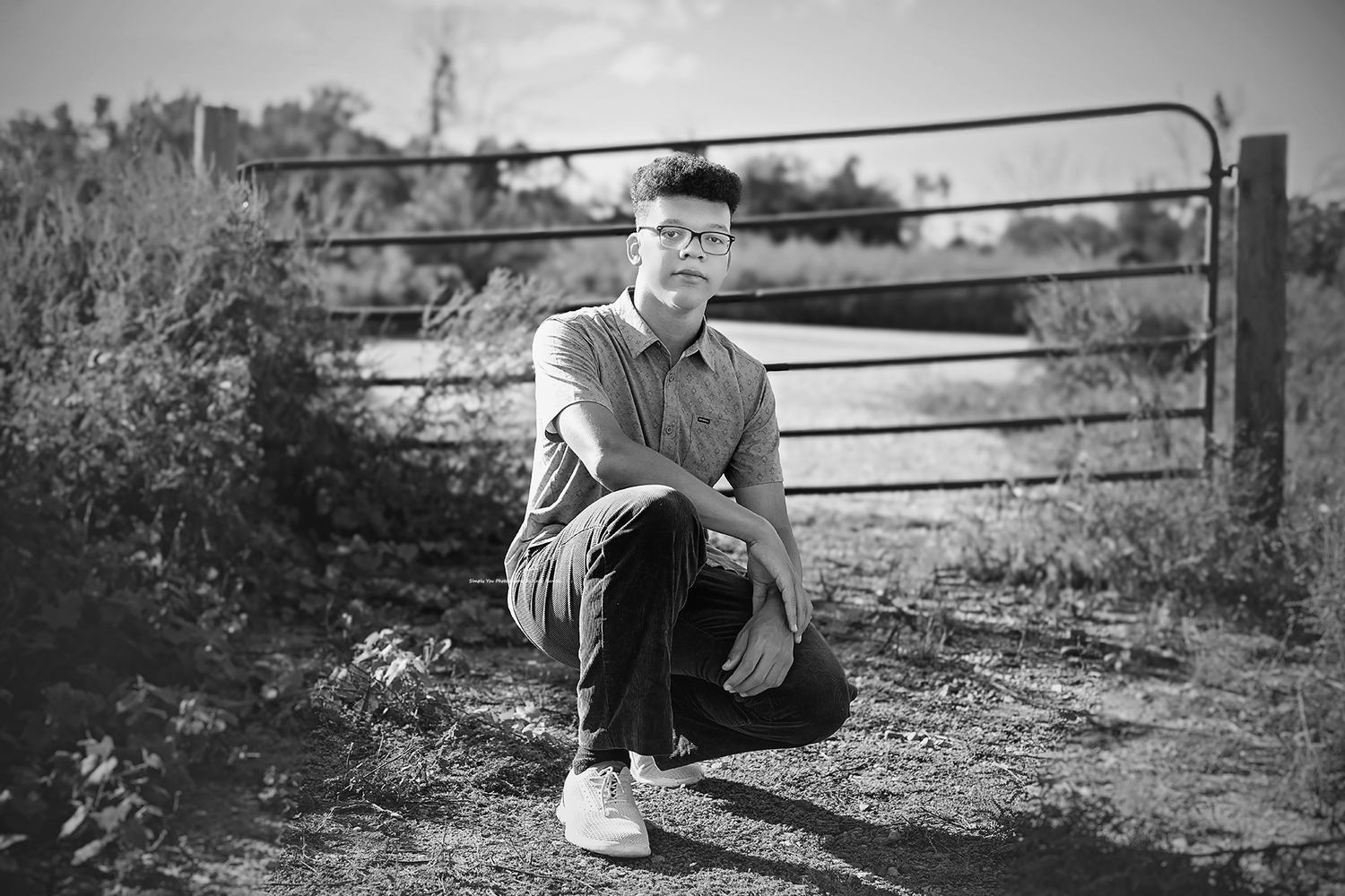 Young man with glasses crouching near a rustic metal gate in a field, black and white portrait photo.