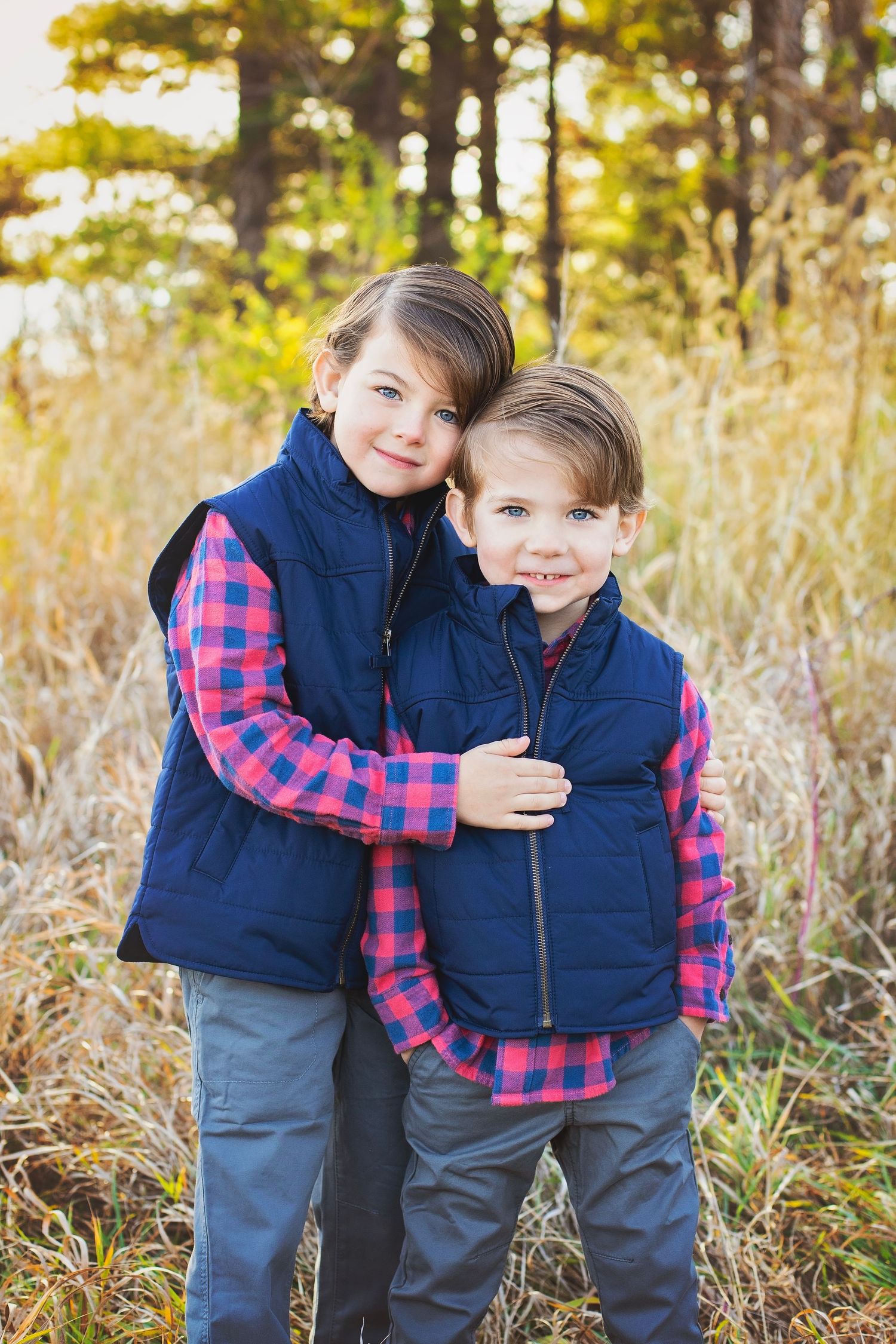Two young brothers wearing matching blue vests and plaid shirts share a warm hug in an autumn field.