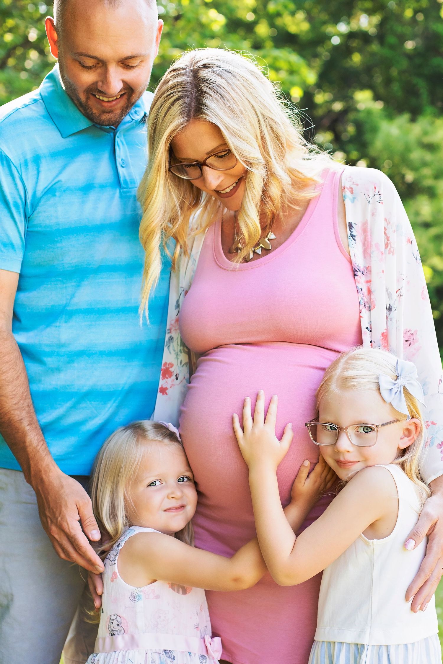 Happy family with pregnant mother in pink dress, father, and two blonde daughters in a park.