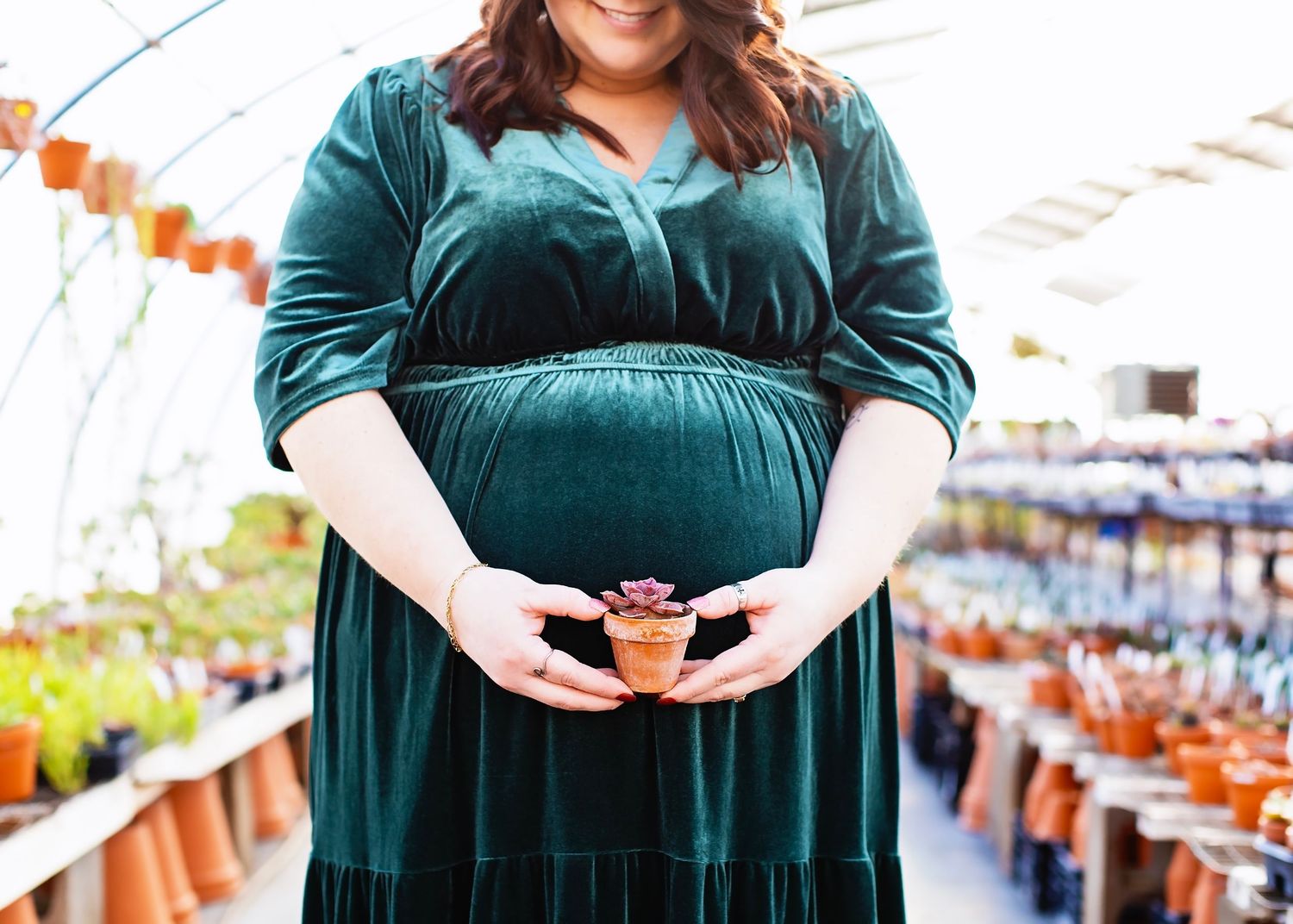 Pregnant woman in emerald green velvet dress holds a small potted plant in a greenhouse nursery.