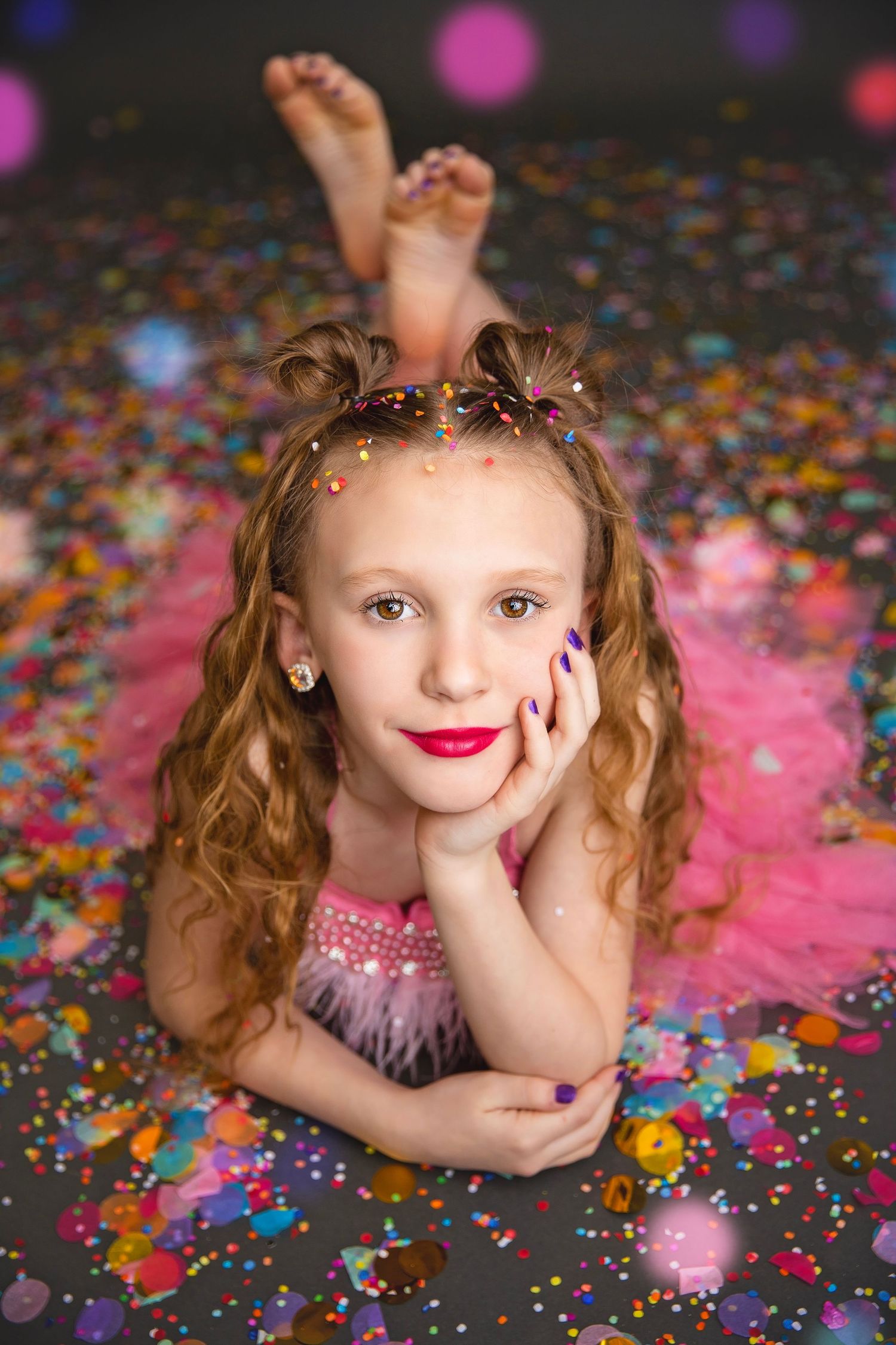 Curly-haired girl in pink tutu lying on confetti-covered floor with feet up.