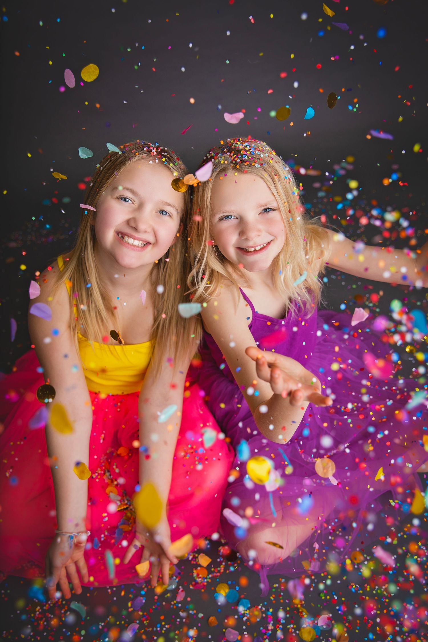 Two smiling girls in bright dresses laughing together as colorful confetti falls around them.