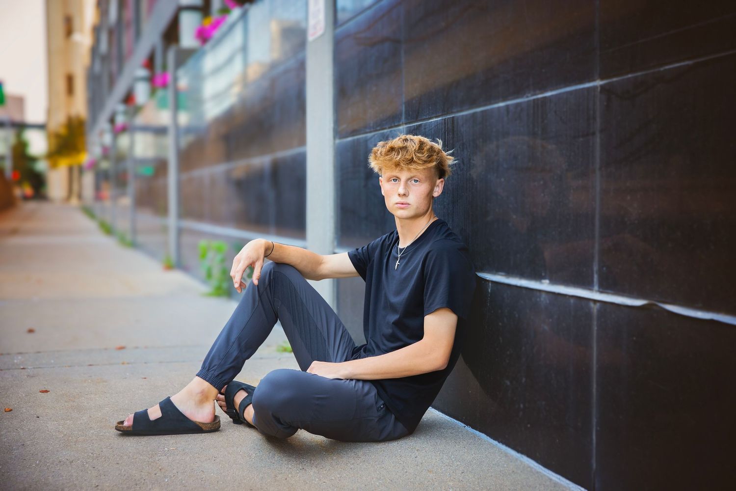 Teen boy with curly blonde hair sits casually against dark tiled wall on urban city sidewalk.