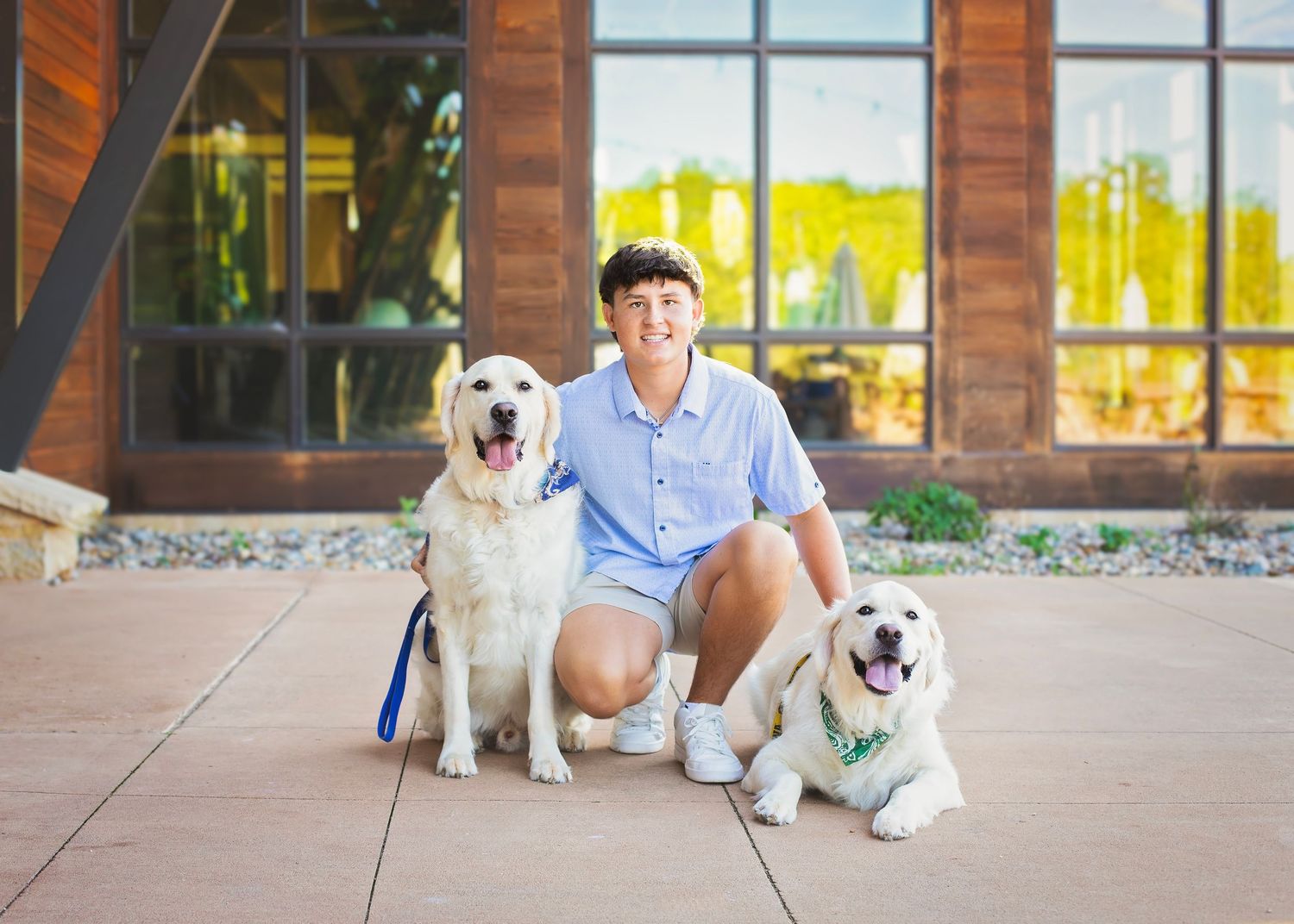 Smiling teen posing with two golden Labrador dogs outside a modern wooden building on a sunny day.