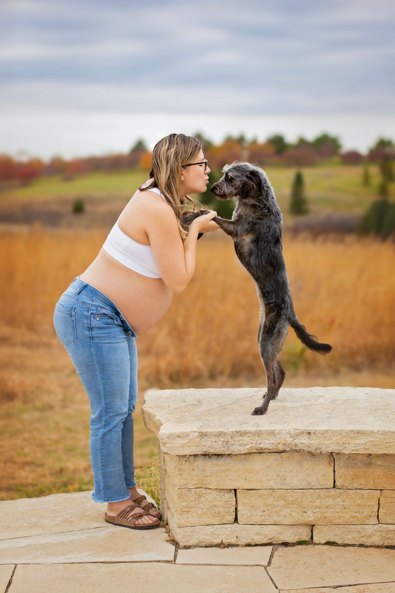 Pregnant woman in white crop top leans in for a kiss from her jumping black dog on stone wall.