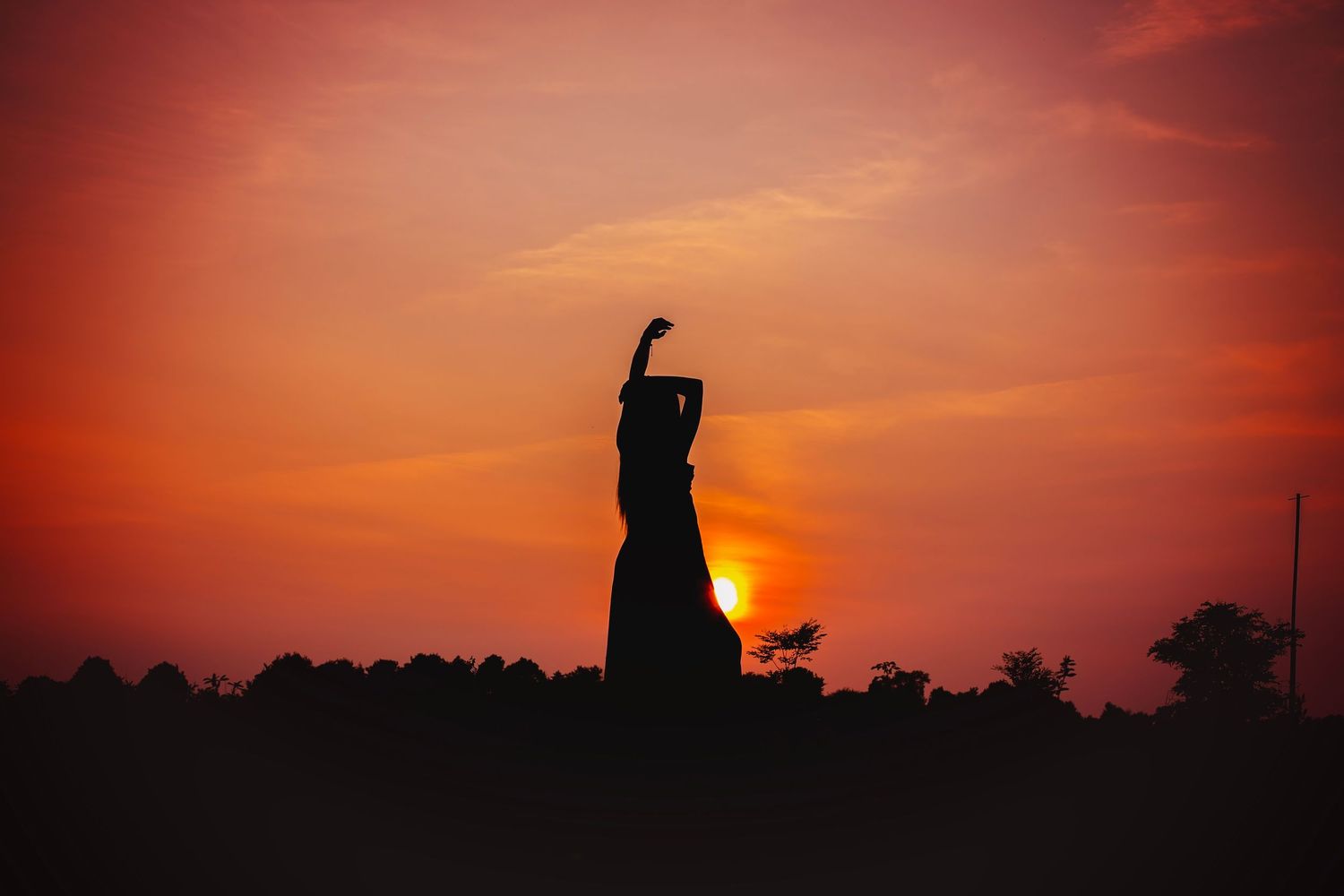 Silhouette of a woman with arms raised against a vibrant orange and red sunset sky.