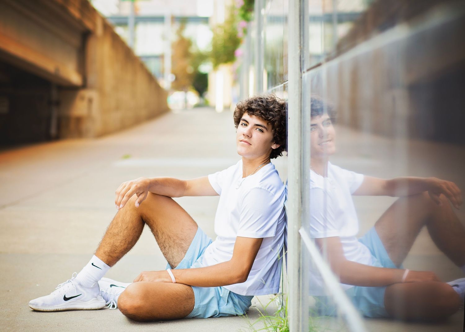 Teen boy in white tee and blue shorts sits against a reflective glass wall outdoors.