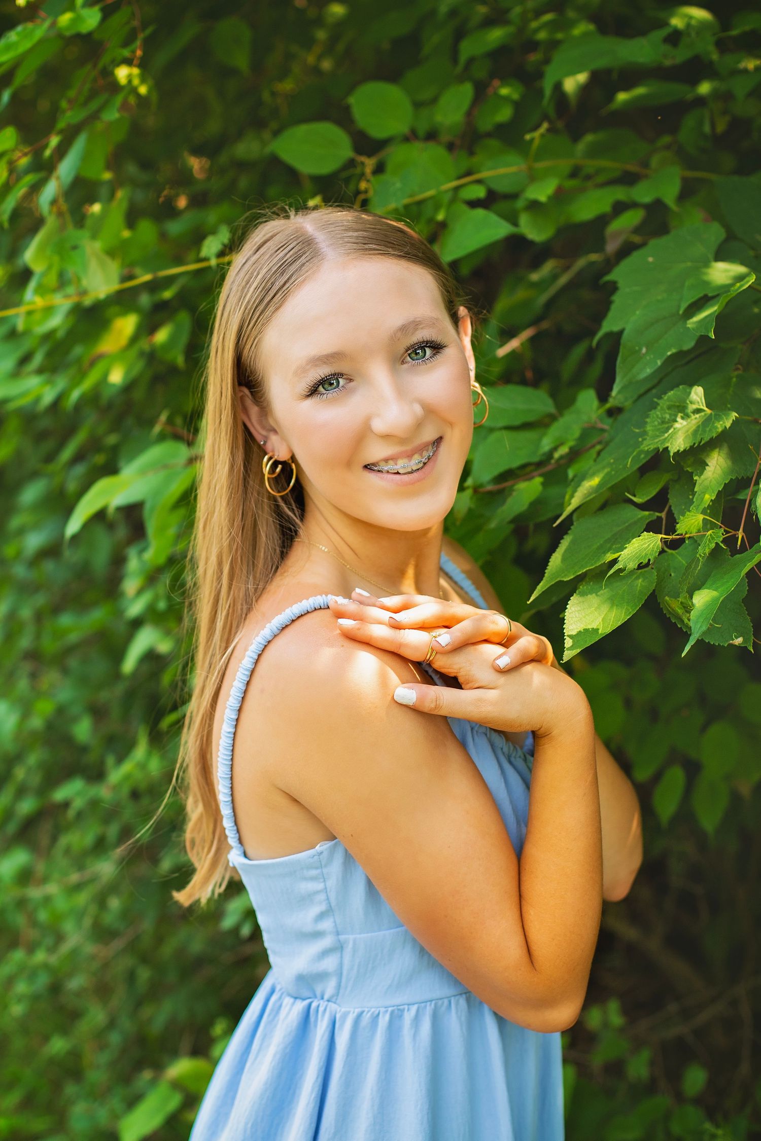 Smiling blonde teen girl in light blue sundress with braces posing against green leafy background.