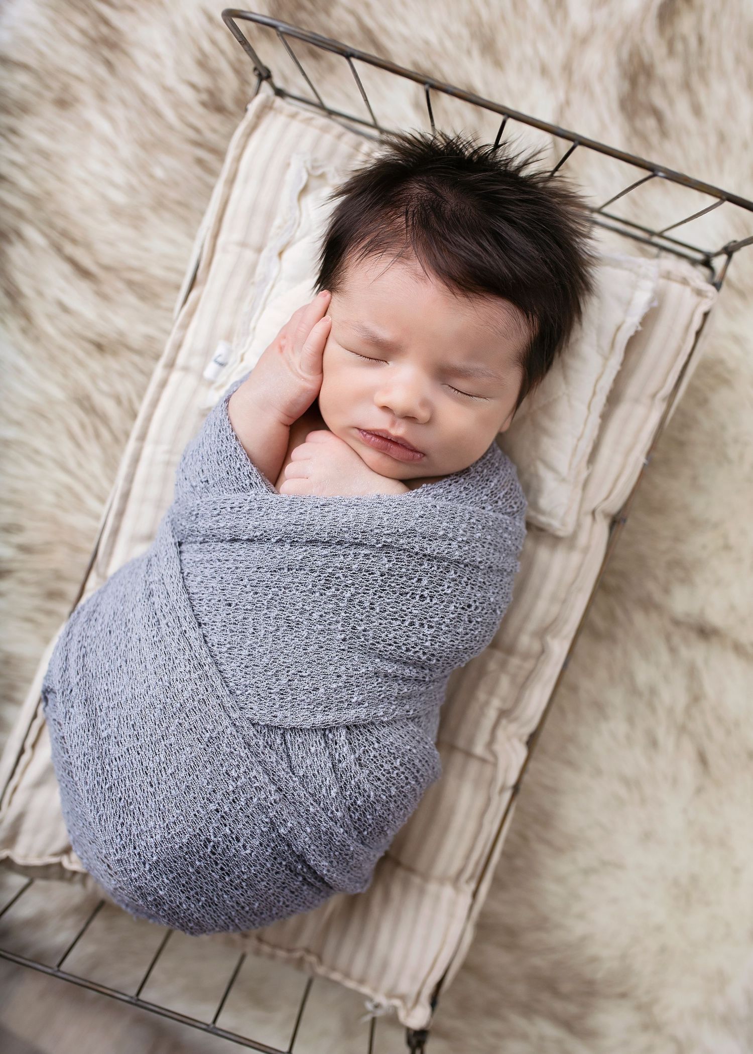 Sleeping newborn baby swaddled in gray knit wrap lying in a small wire basket with fur blanket.