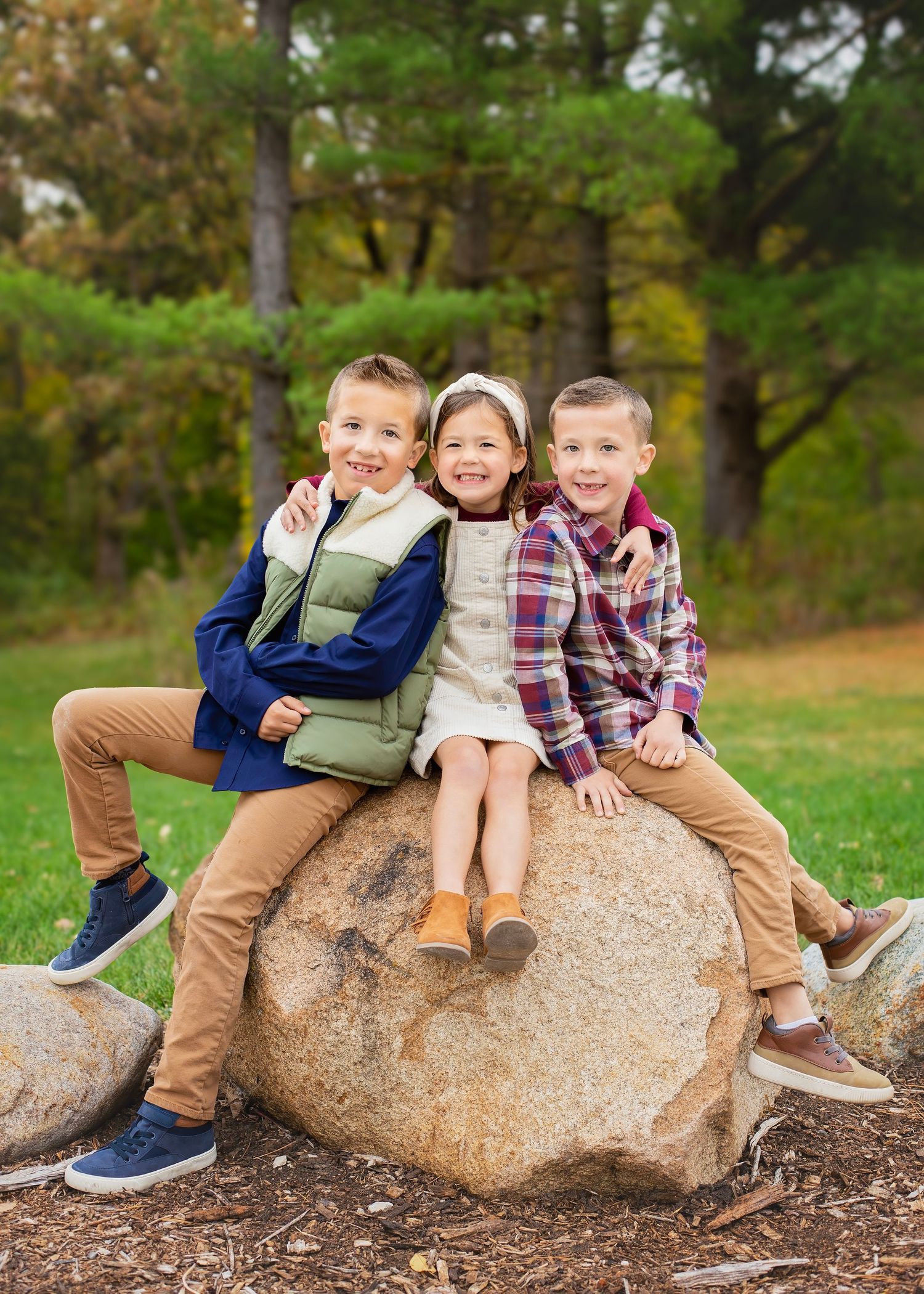Three young children sitting on a large boulder in a park during autumn.