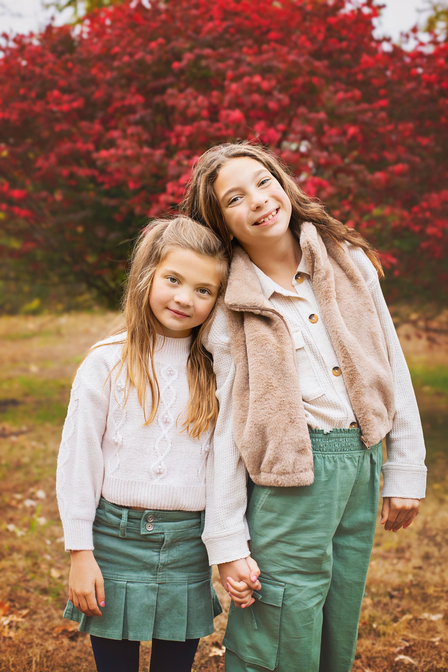 Two young girls in green skirts hold hands and smile together in front of vibrant red autumn foliage.