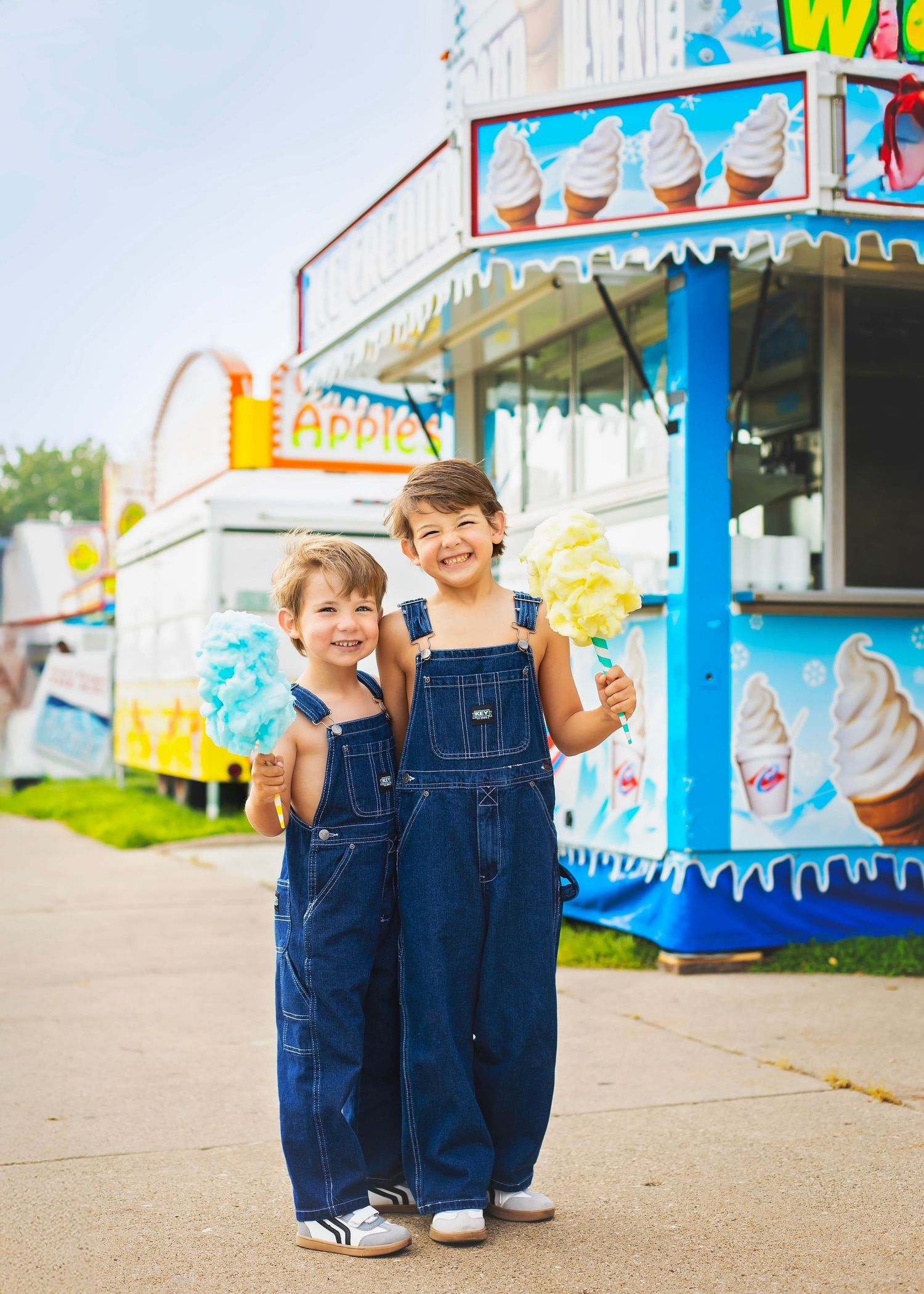 Two smiling boys in denim overalls holding cotton candy treats at a summer fair food stand.