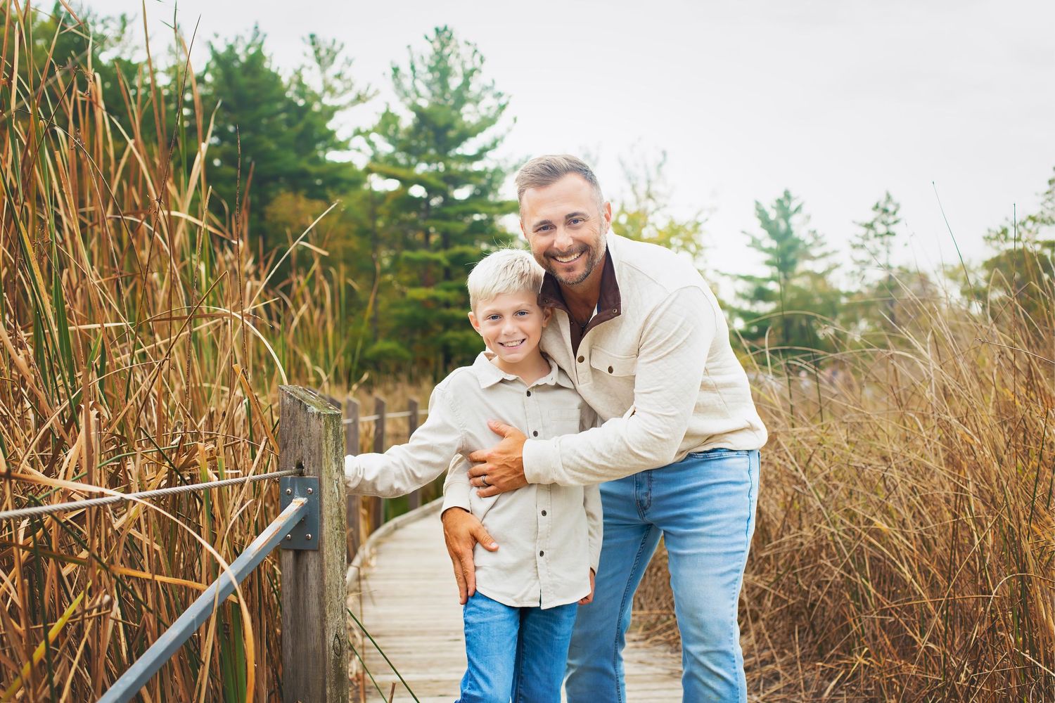 A smiling father and young son pose together on a wooden boardwalk surrounded by tall golden reeds in autumn.
