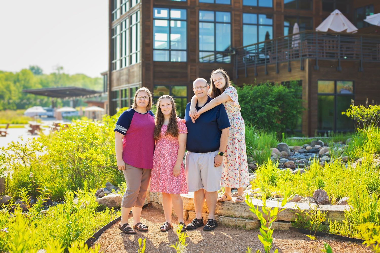Family of four posing together outdoors near modern building with lush green garden landscape.