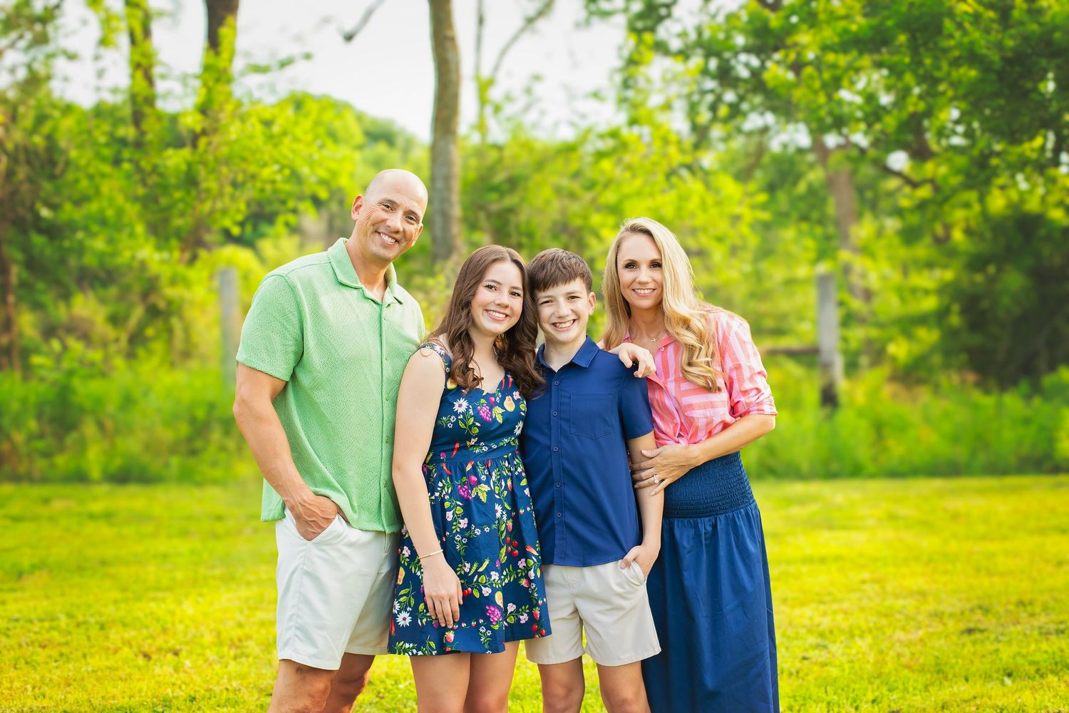 Family of four smiling together on green grass with trees in background during outdoor photo session.