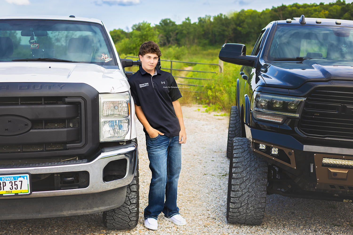 Young man standing between two large pickup trucks on a gravel path with green fields in the background.