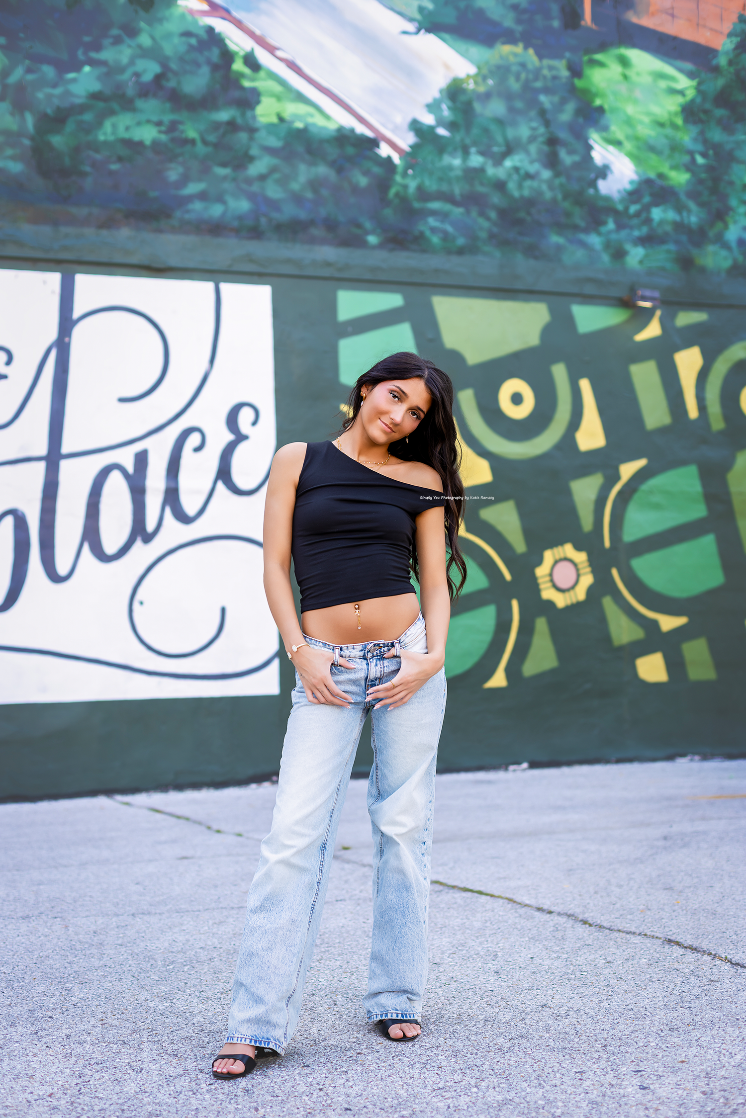 Young woman in black one-shoulder crop top and light wash flare jeans posing in front of colorful mural.
