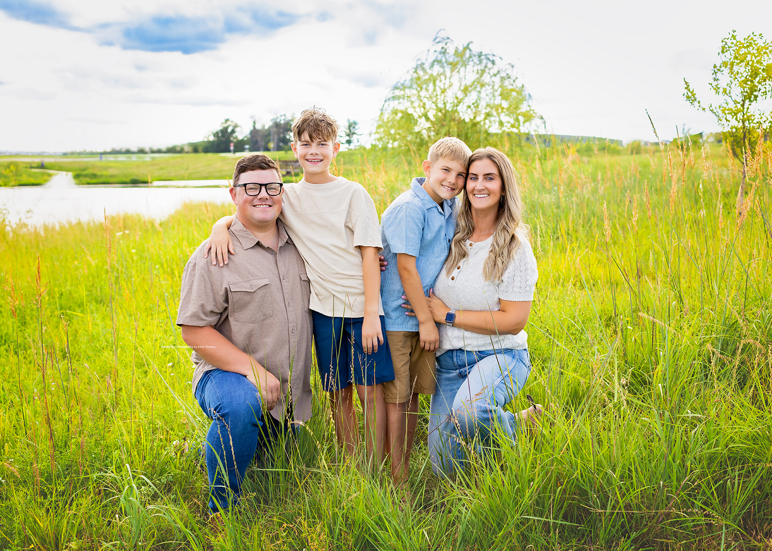 Family of four posing in a lush green field with a scenic pond and blue sky in the background.