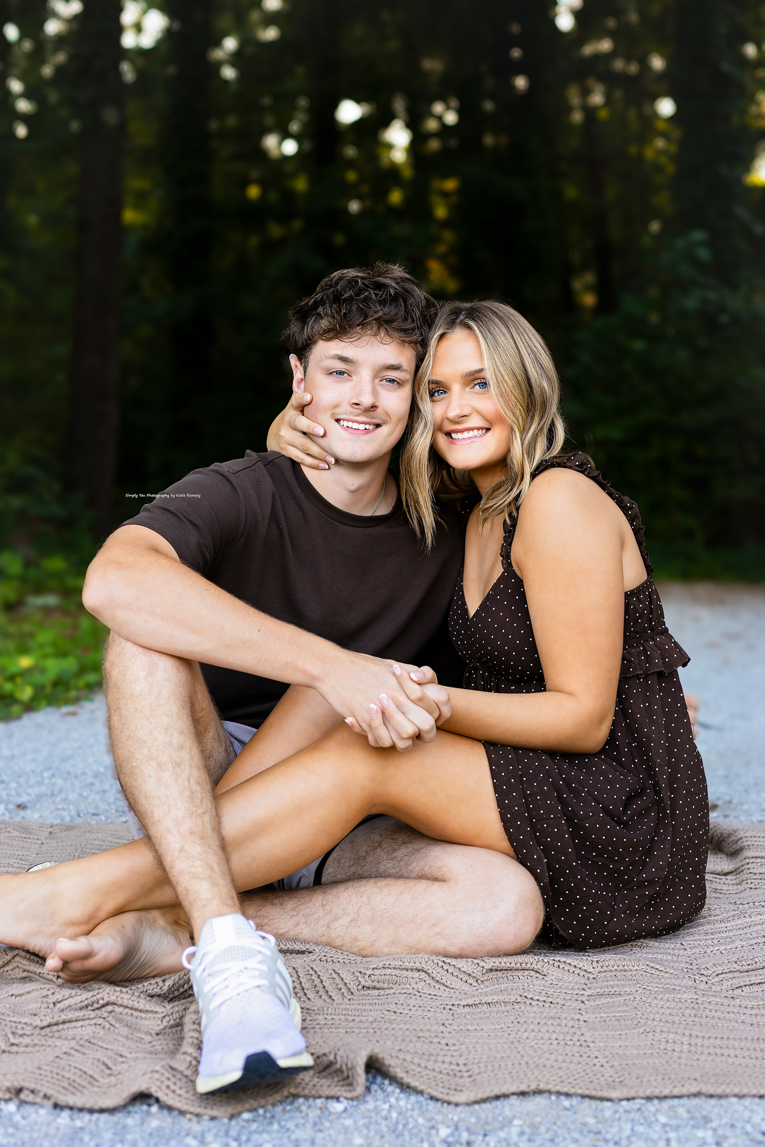 A smiling young couple sits together on a blanket outdoors, holding hands in front of a lush green forest backdrop.