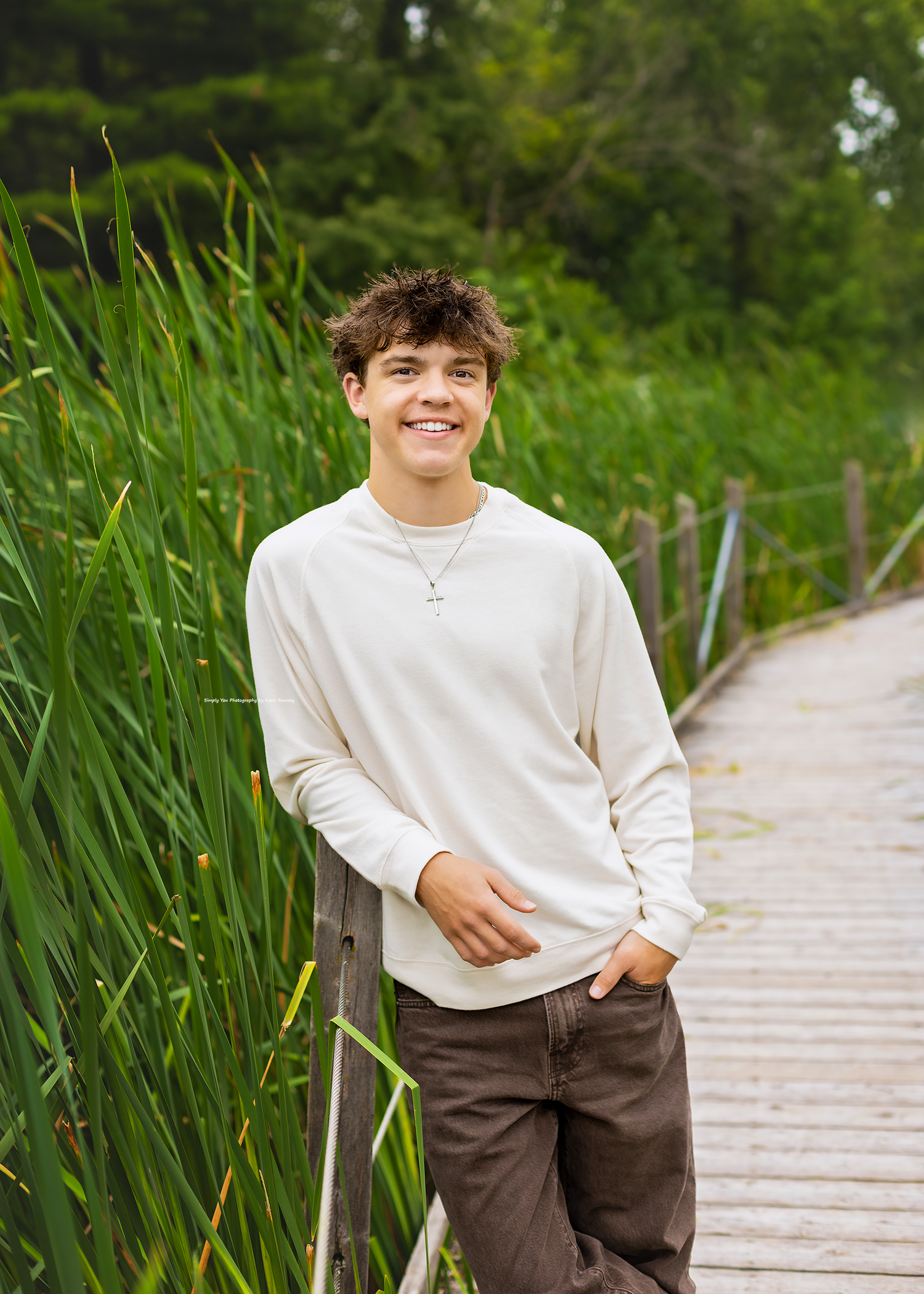 Smiling teen boy in cream sweatshirt and brown pants poses on wooden boardwalk surrounded by tall green reeds.