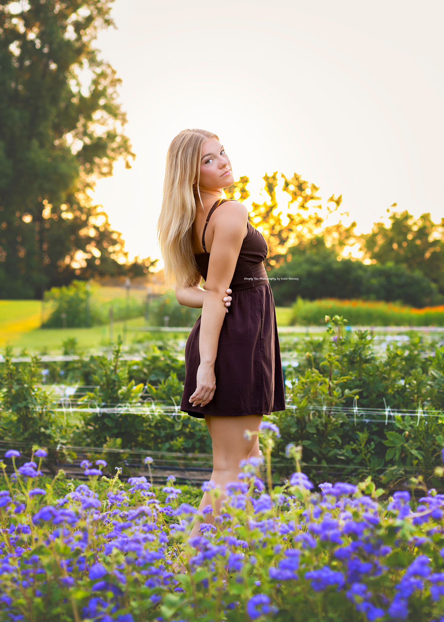 Blonde woman in dark dress standing among purple wildflowers at golden hour sunset in a lush green garden.