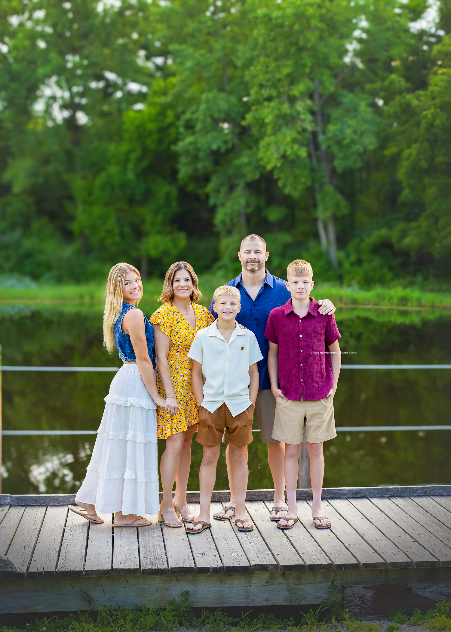 Family of five posing on a wooden dock surrounded by lush green trees and a calm lake backdrop.