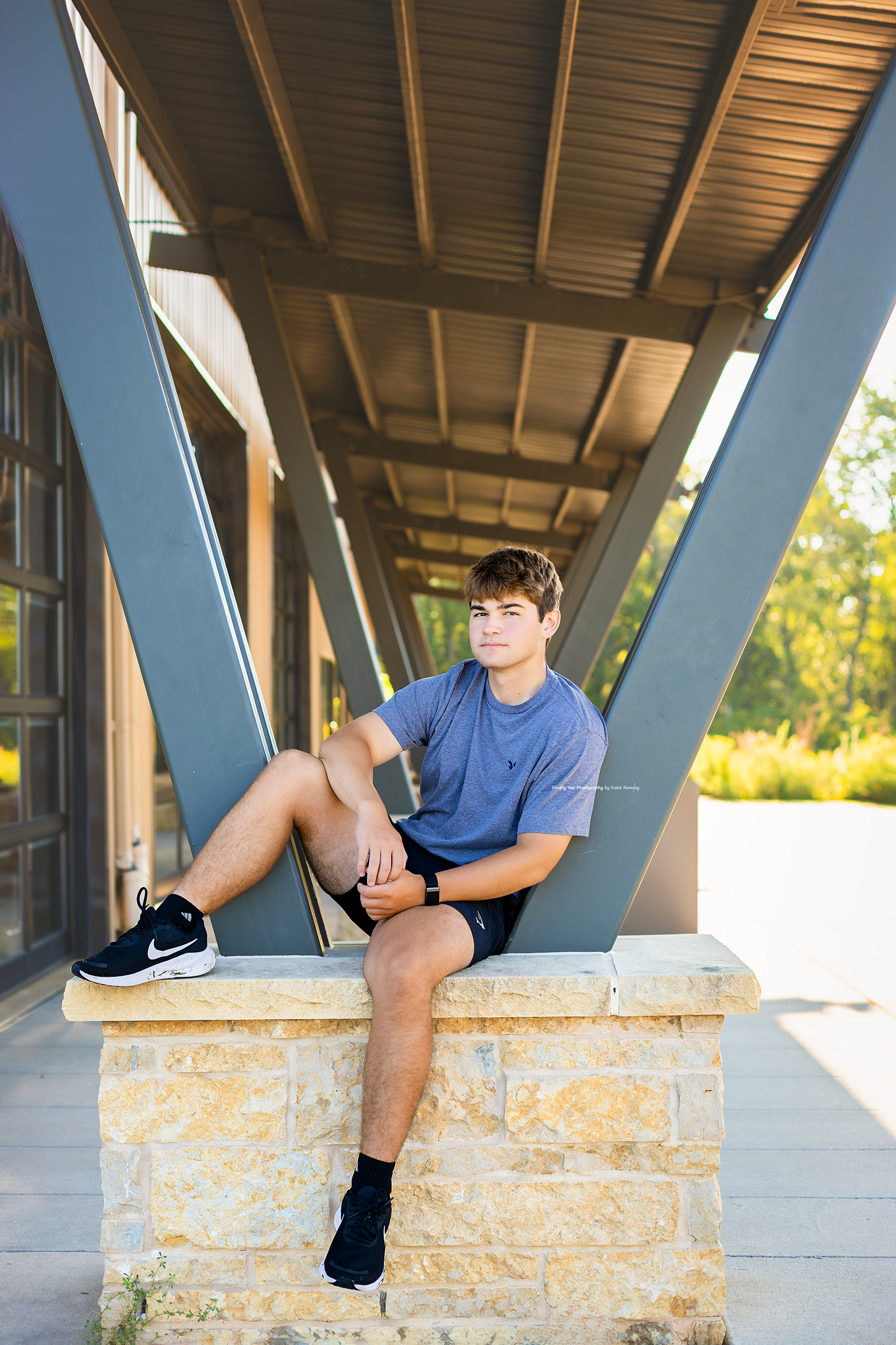 A young man in a blue shirt and shorts sits on a stone ledge under a modern covered walkway with steel beams.