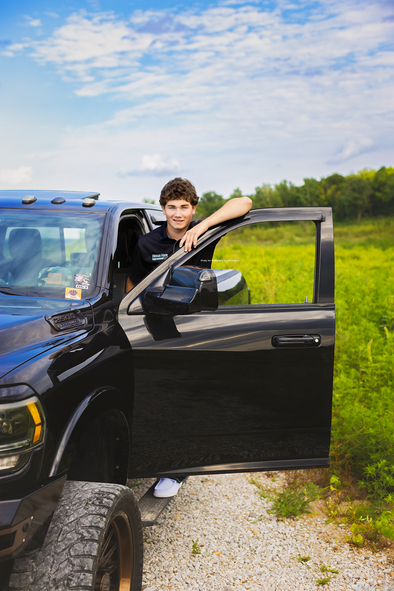 Young man leaning on open door of black pickup truck parked on gravel path with green field and blue sky backdrop.