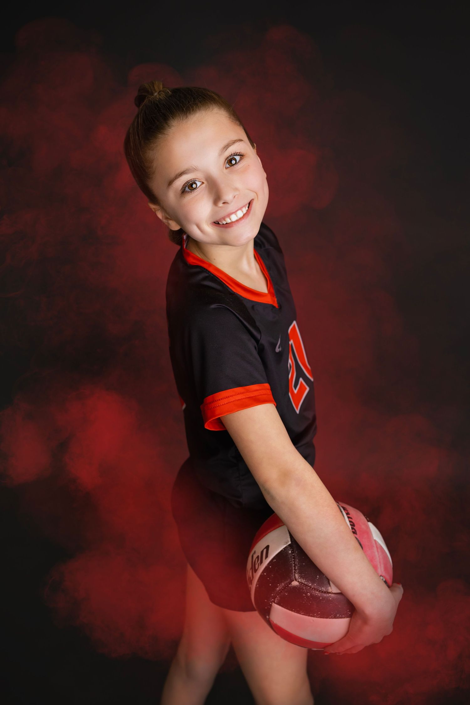 Young smiling girl in black volleyball uniform holding ball turning toward camera against red smoky backdrop.