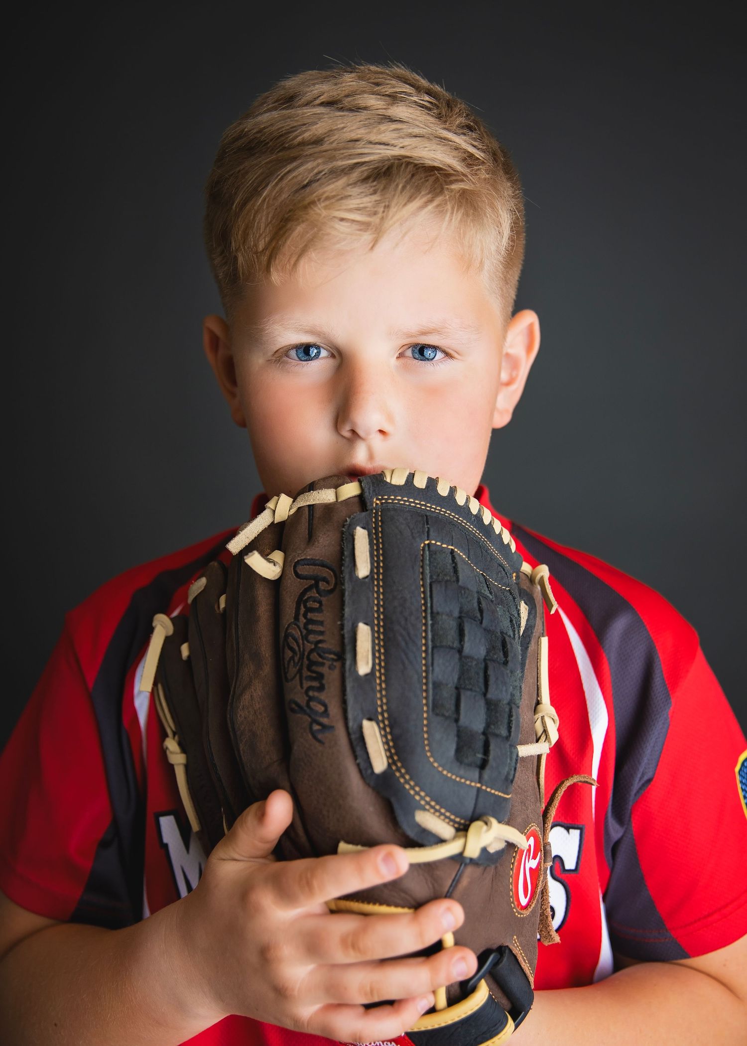 Young blonde baseball player in red Rawlings jersey hides face behind large brown leather baseball glove.
