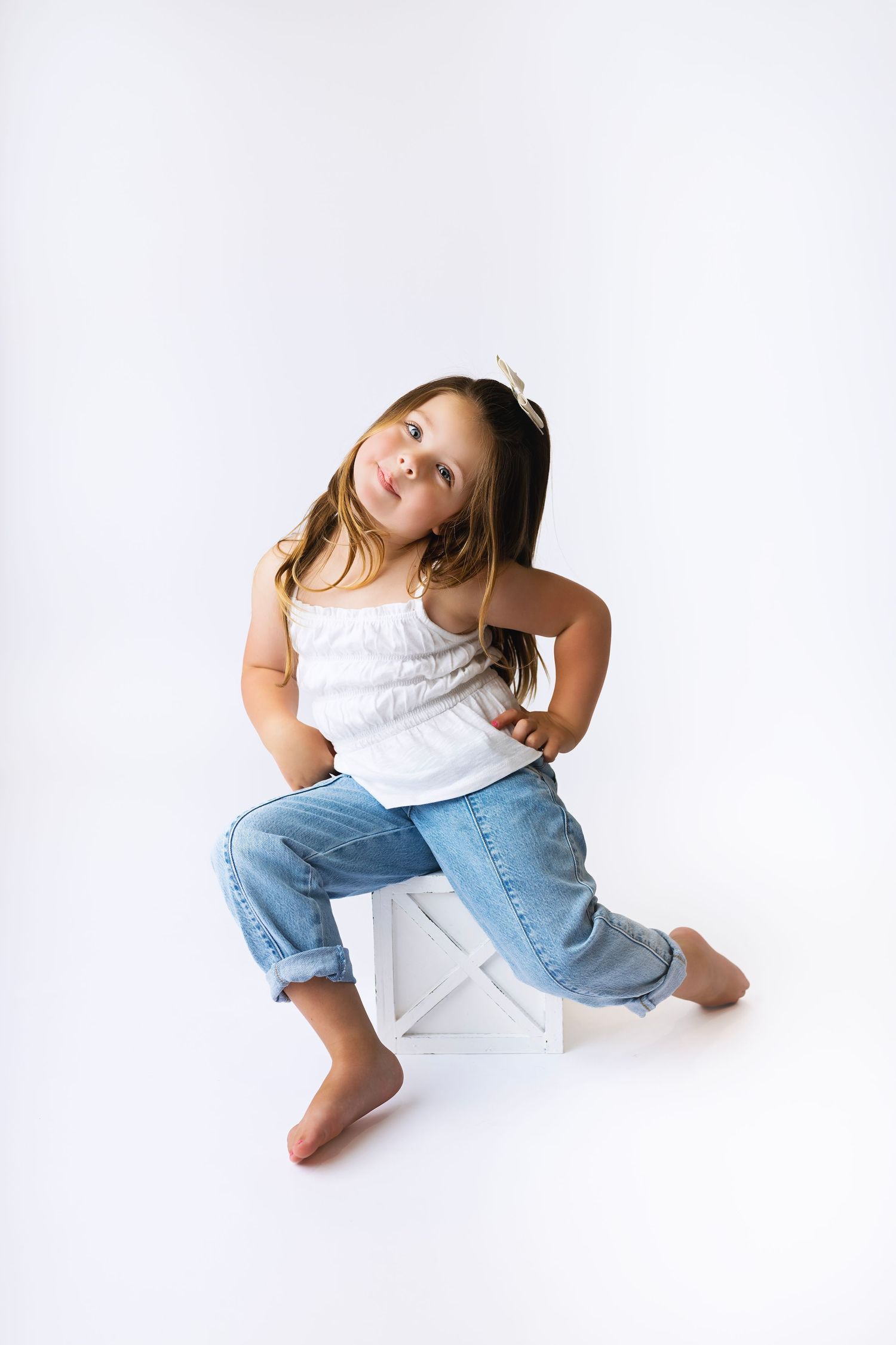 Young girl in white tank top and jeans sitting on white box, posing confidently with hand on hip.