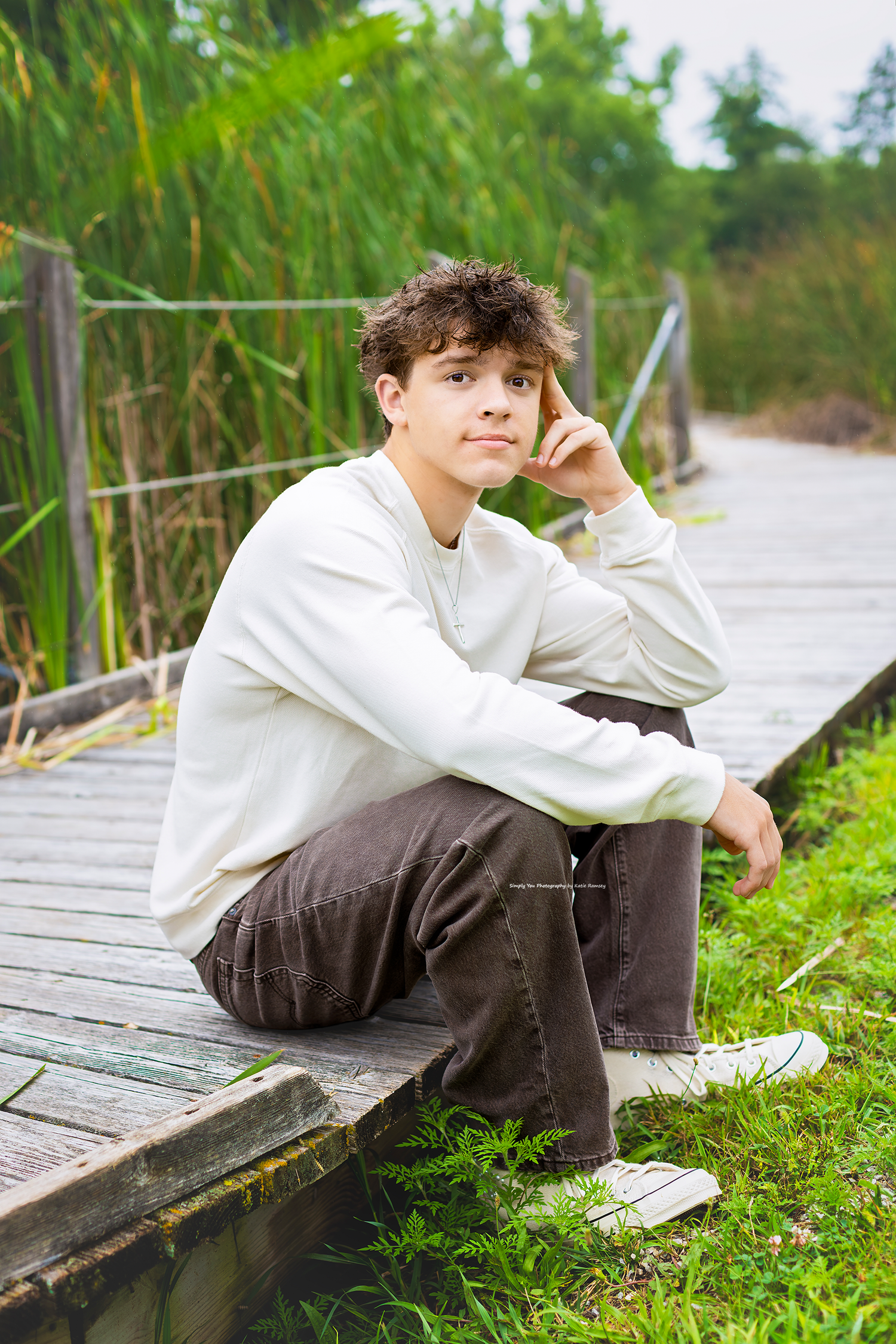 Teen boy in white sweatshirt and brown jeans sitting on wooden boardwalk surrounded by lush green reeds.