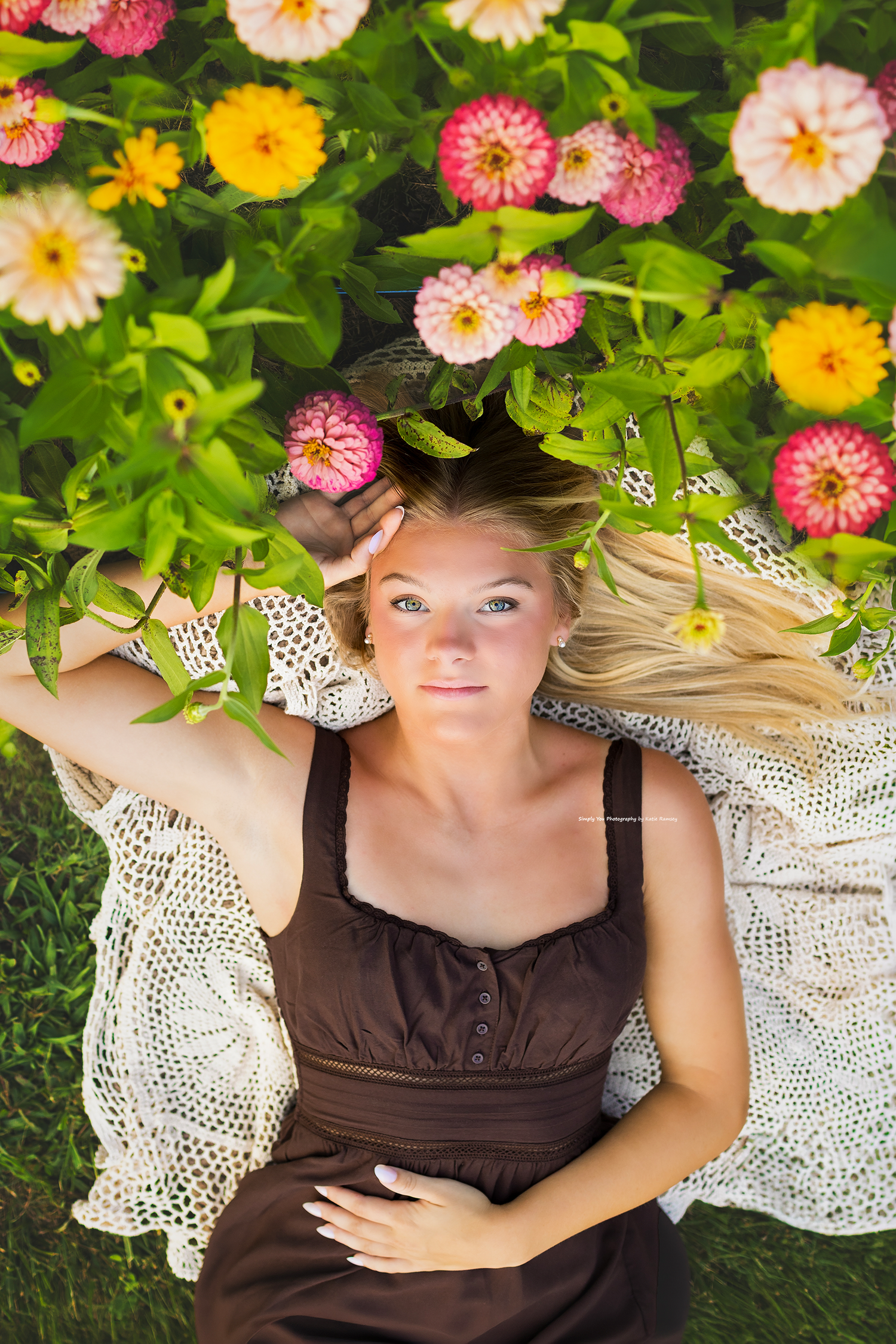 Young blonde woman lying in a garden surrounded by colorful zinnia flowers, wearing a brown dress.