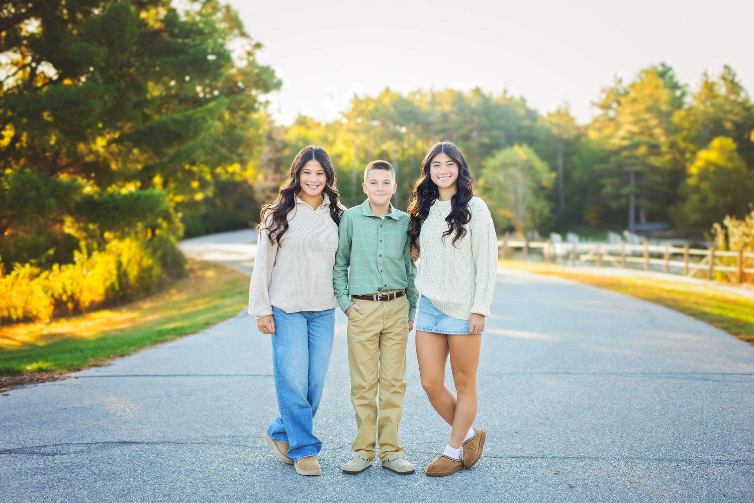 Three siblings standing together on a sunlit road with autumn trees in background.