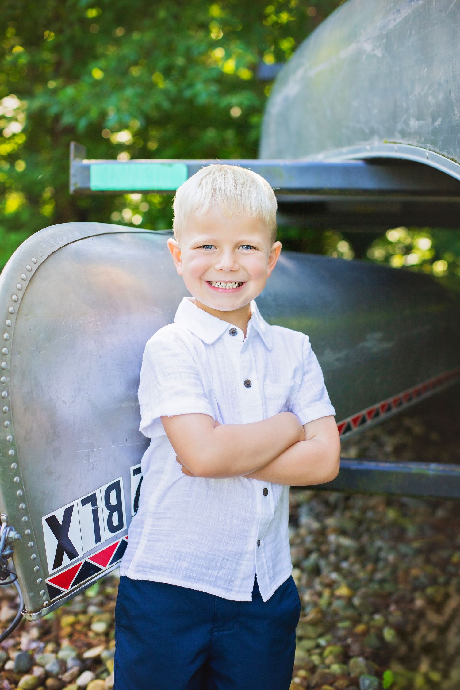 Smiling blonde boy in white shirt stands arms crossed in front of a vintage boat outdoors.
