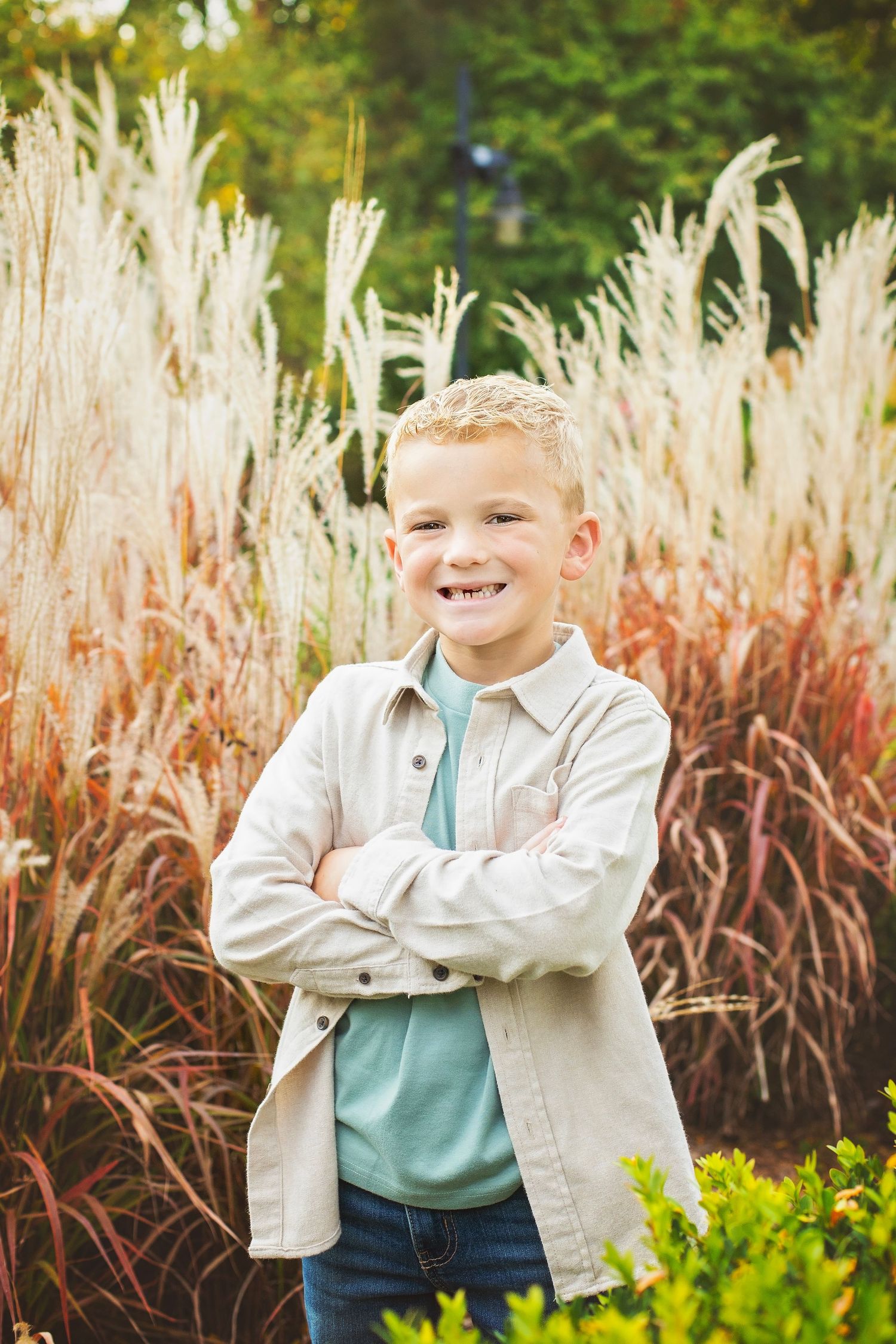 A smiling blonde boy stands with arms crossed in front of ornamental grasses.