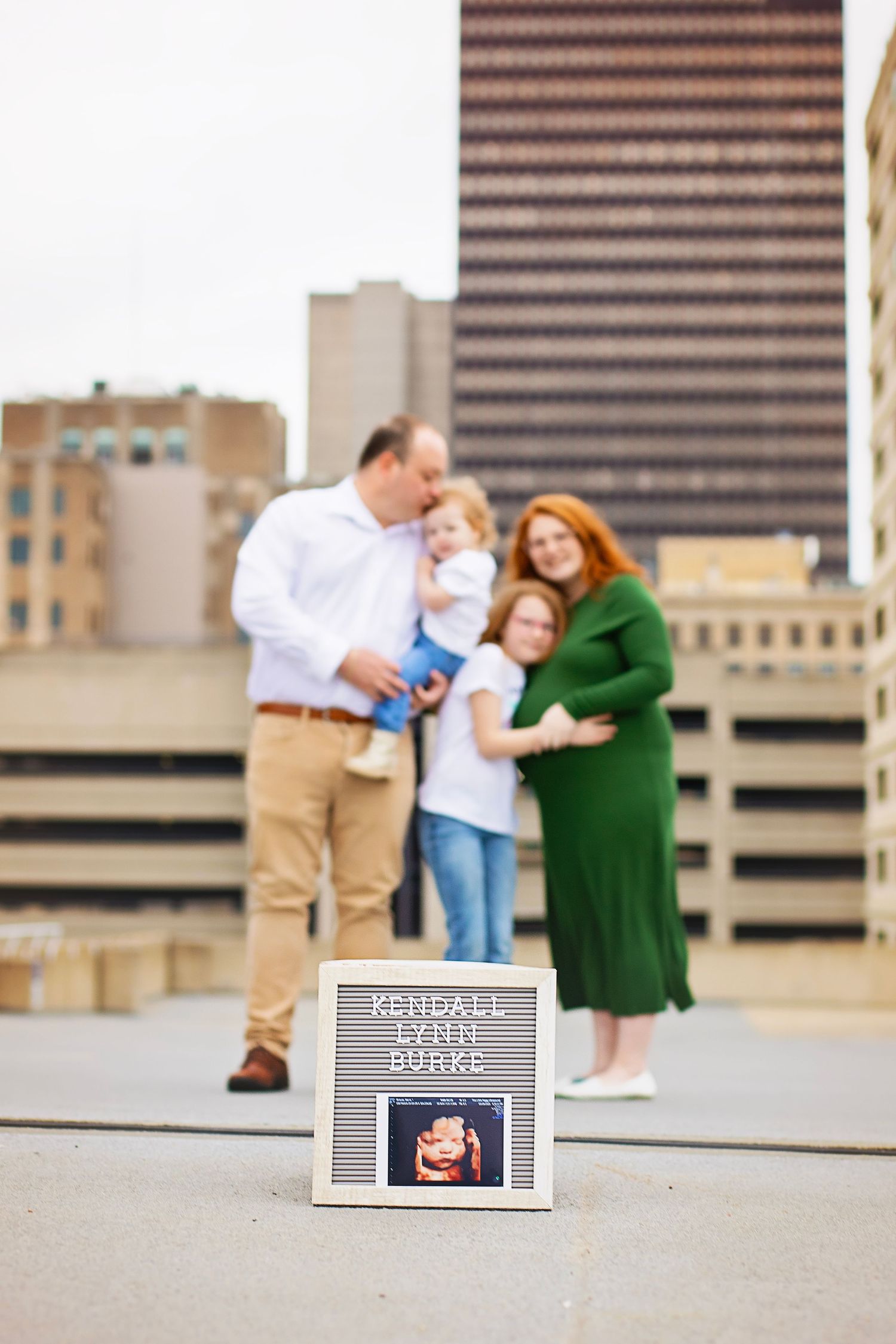 Family maternity photo with Kendall Lynn Burke sonogram letterboard on urban rooftop parking deck.