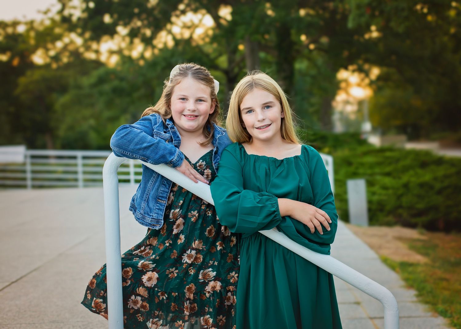 Two young sisters leaning on a white railing outdoors, smiling, wearing a floral dress and a green dress.