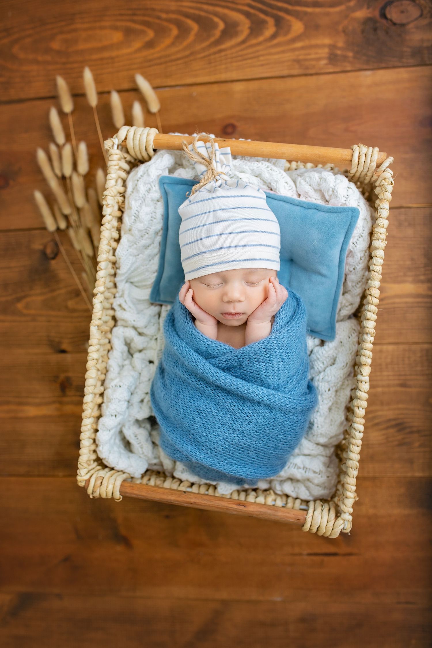 Sleeping newborn baby wrapped in blue knit swaddle in a wicker basket with tiny pillows.