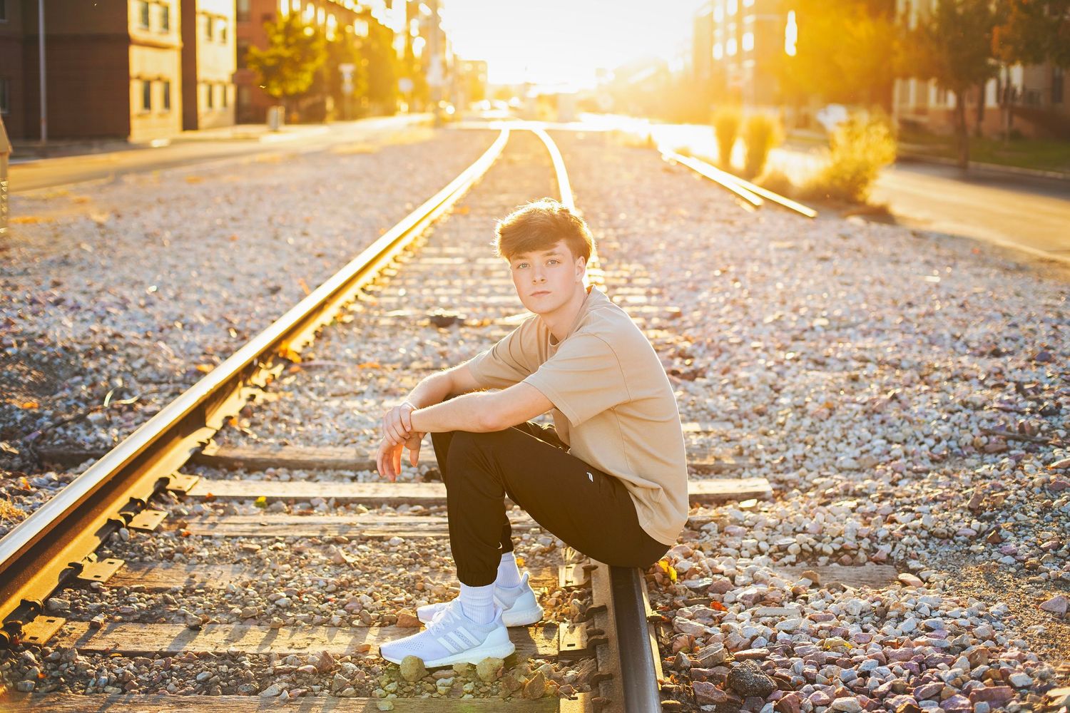 Teen boy in beige outfit sitting on railroad tracks during golden hour sunset.